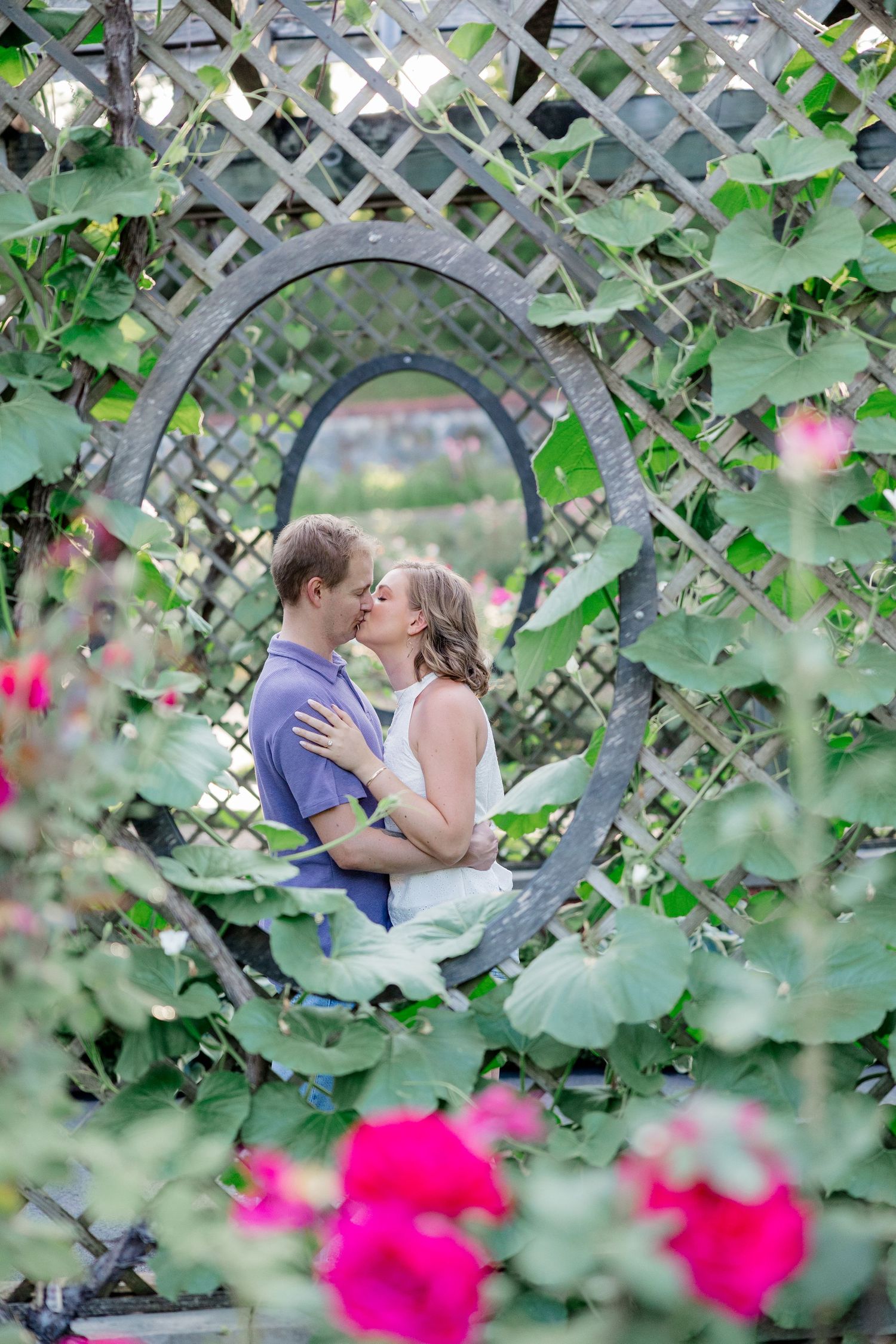 A couple embraces behind a lattice trellis surrounded by vibrant pink flowers and lush greenery.