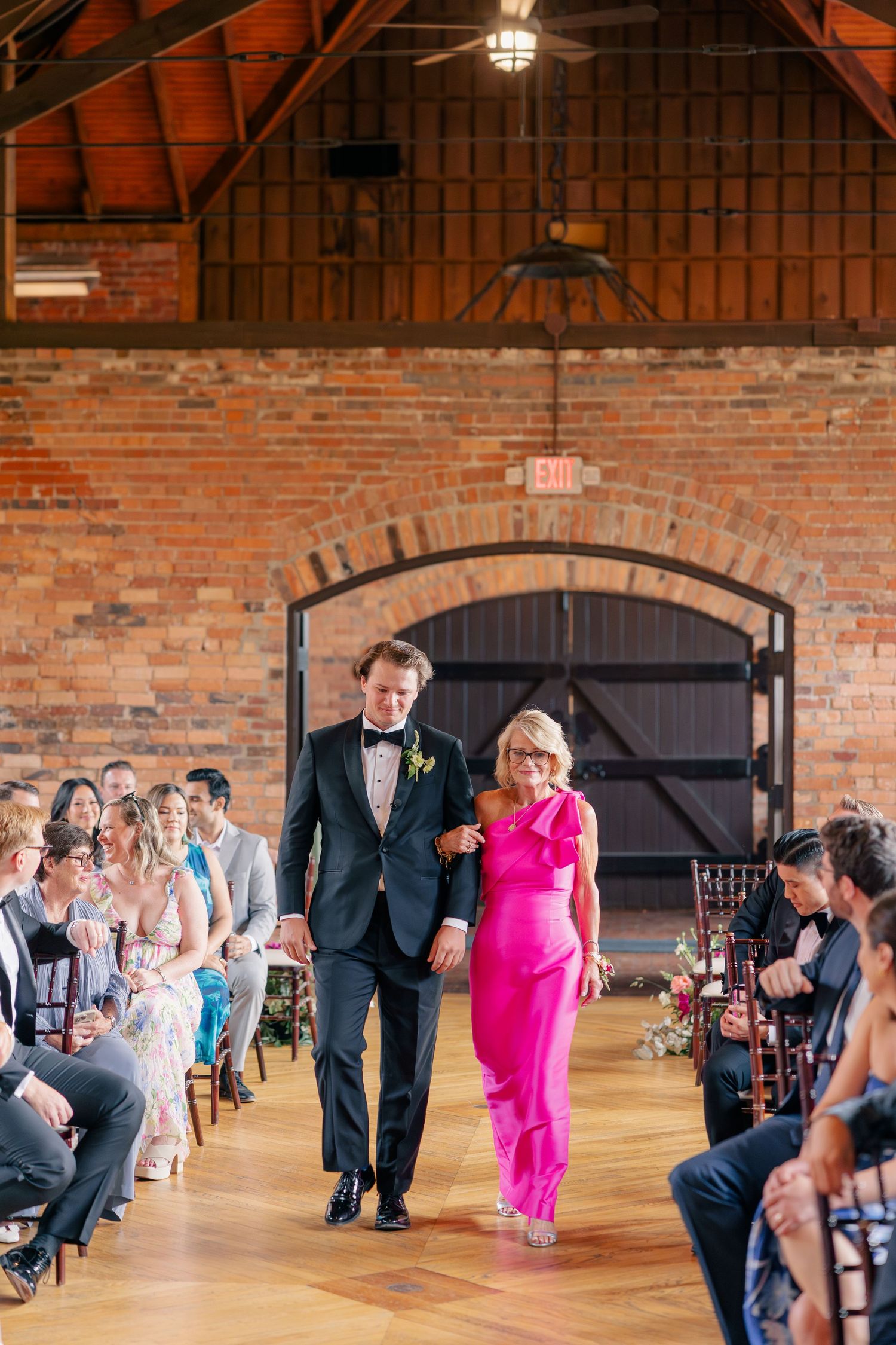 Groom in black tuxedo escorts woman in hot pink gown down wedding ceremony aisle in brick venue.