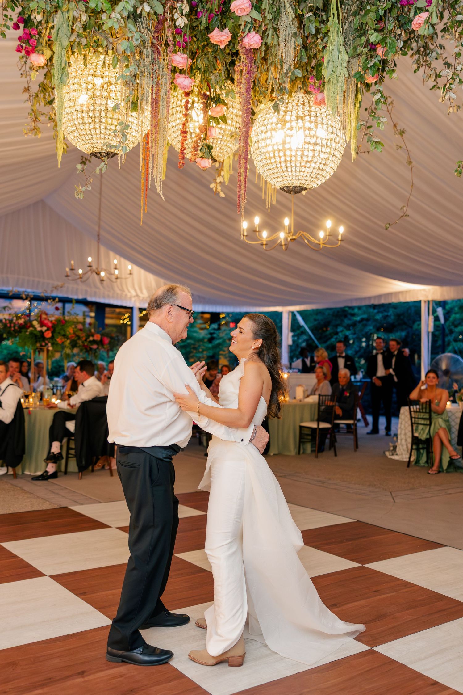 Bride dances with father under floral chandeliers at elegant tented wedding reception.