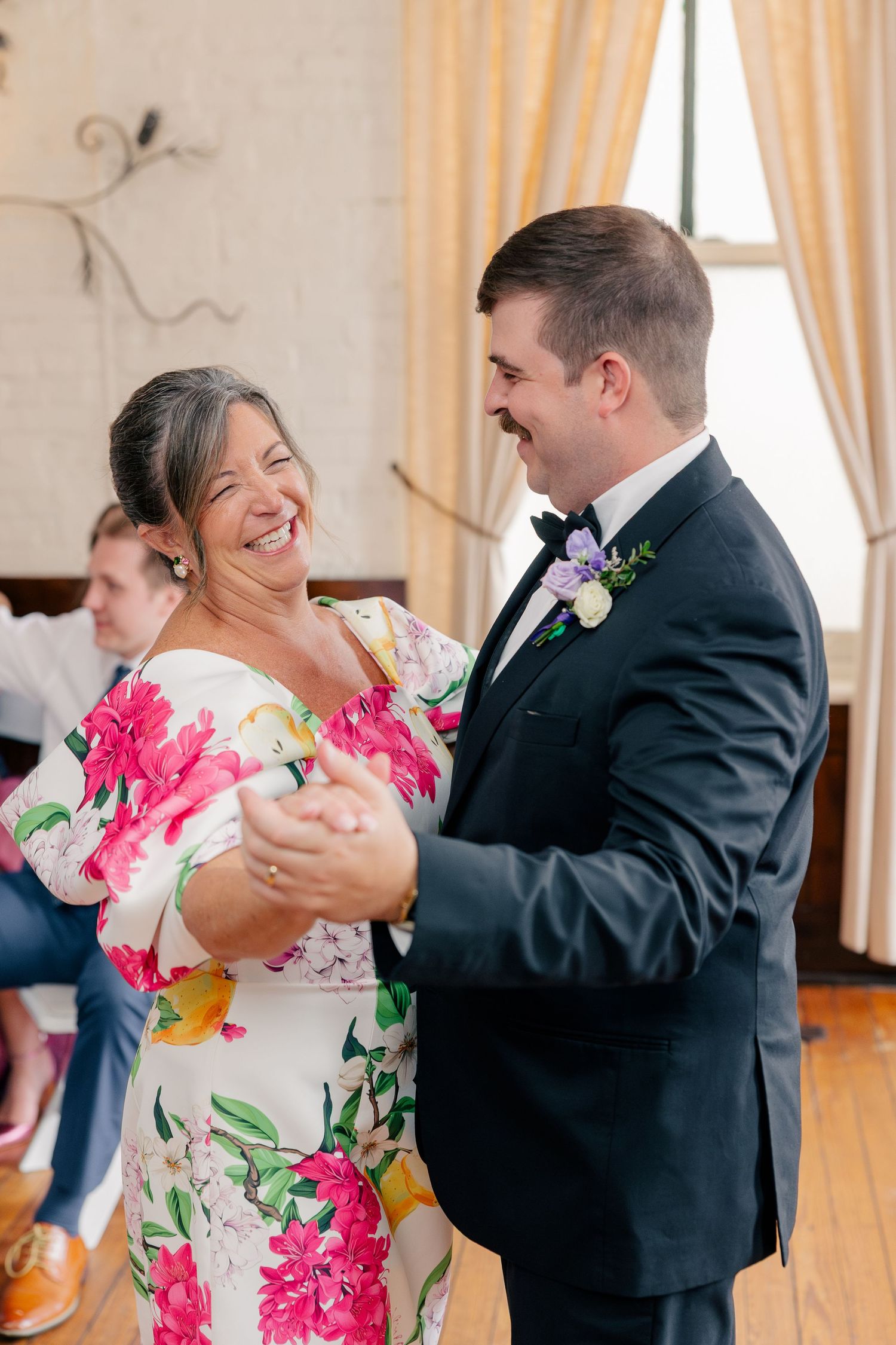 Groom in black tuxedo shares a joyful mother-son dance with woman in floral dress at wedding reception.