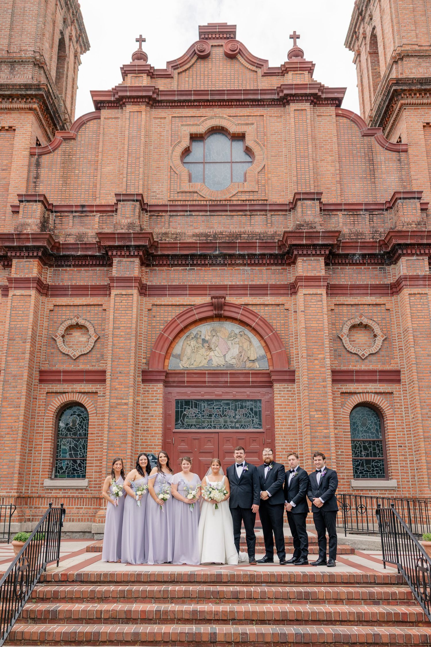 Wedding party poses on steps of ornate red brick church with bridesmaids in lavender.