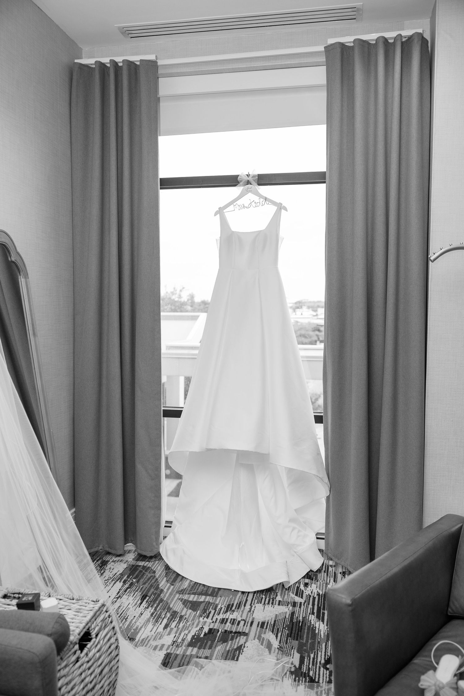 Black and white photo of elegant white wedding gown hanging in hotel window before the ceremony.