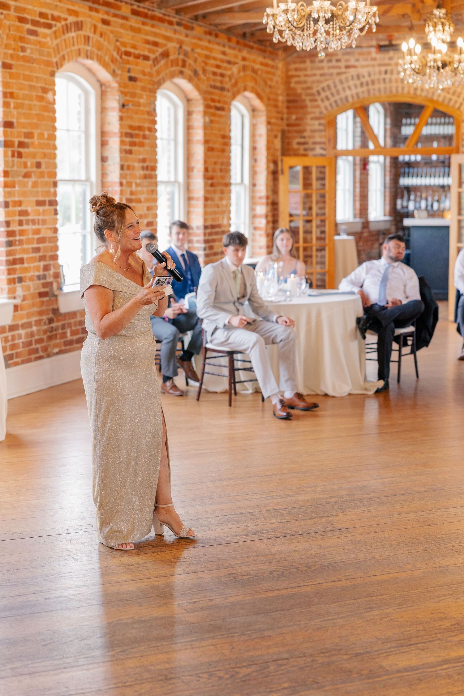 Elegantly dressed woman delivers a heartfelt wedding toast speech in a grand rustic brick ballroom with chandeliers.