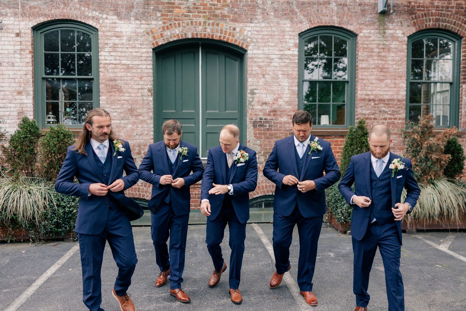 Five groomsmen in matching navy suits adjust jackets outside rustic brick building with green doors.