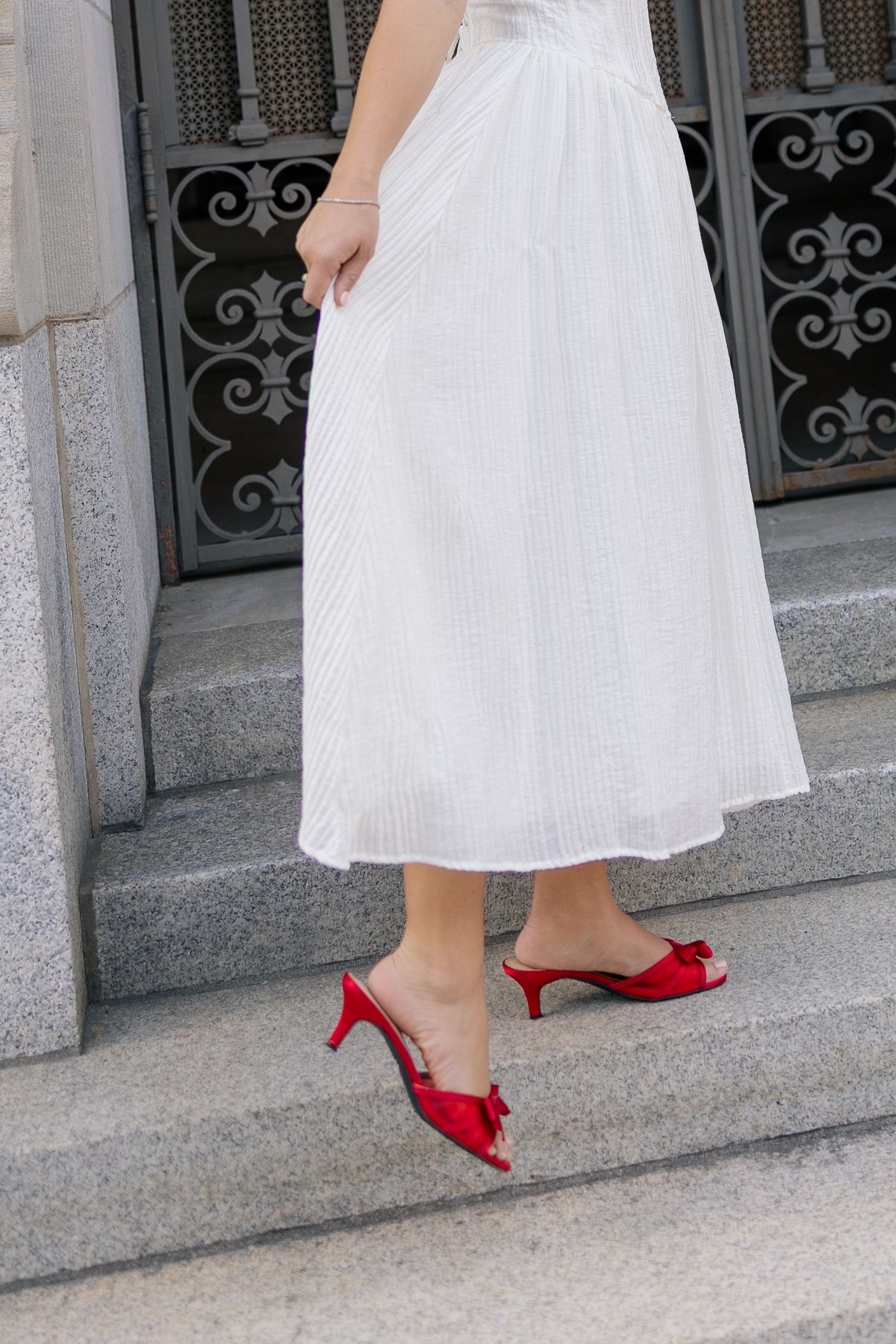 Woman in white midi skirt and red kitten heel mules with bow detail standing on stone steps.