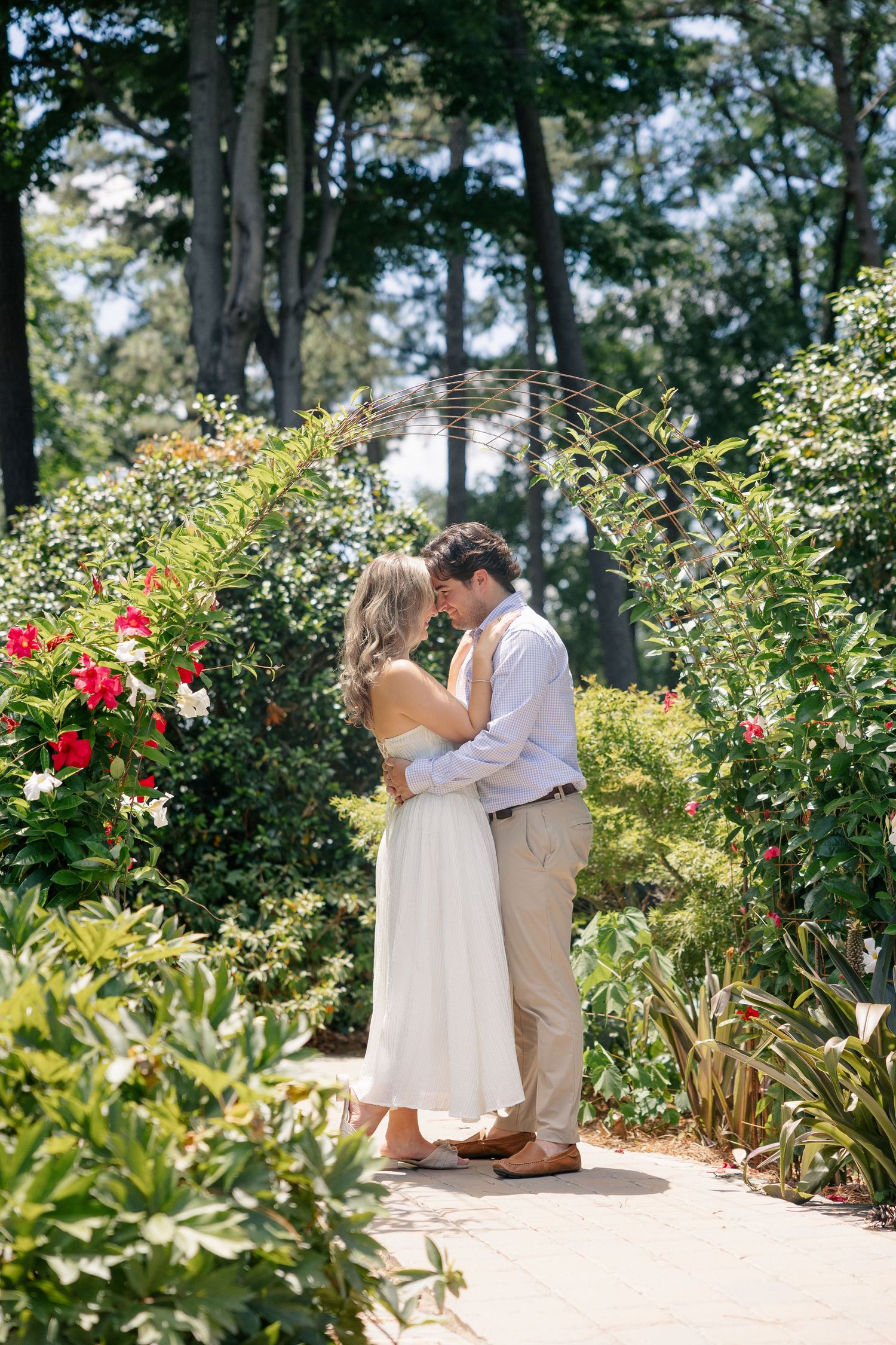 Couple embracing in a lush garden pathway surrounded by blooming red and white flowers during engagement session.