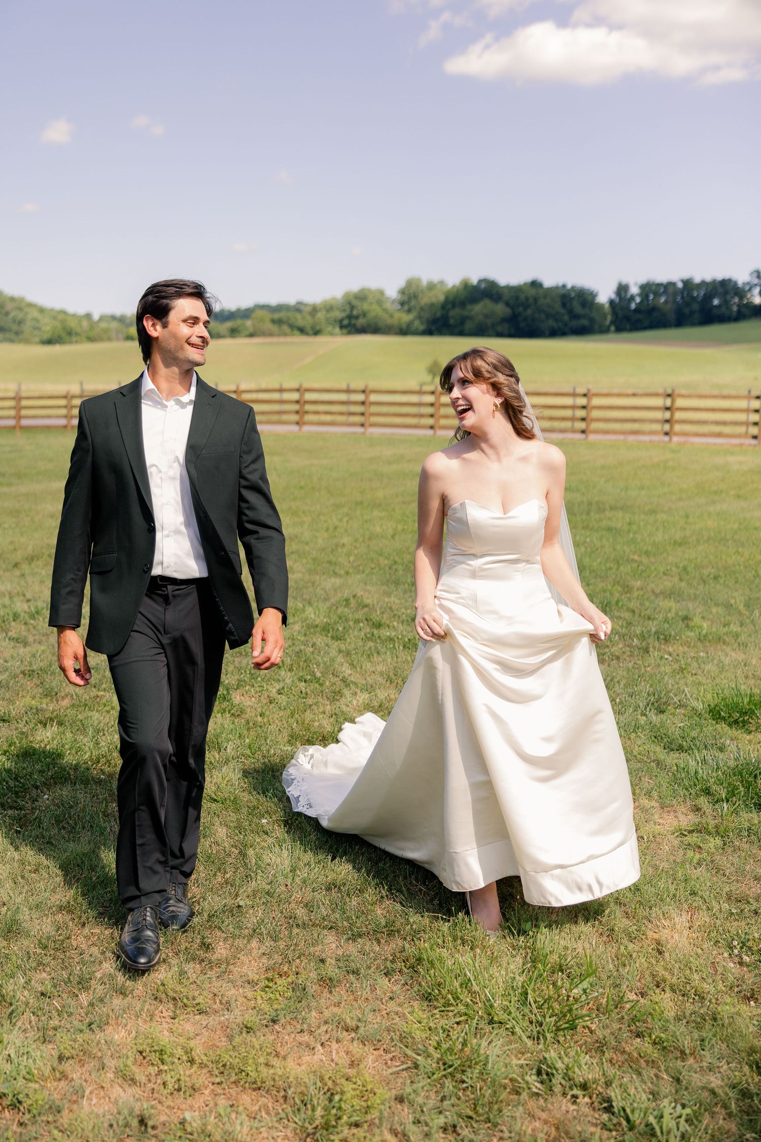 Happy bride and groom walking hand in hand through a green field with a wooden fence backdrop.
