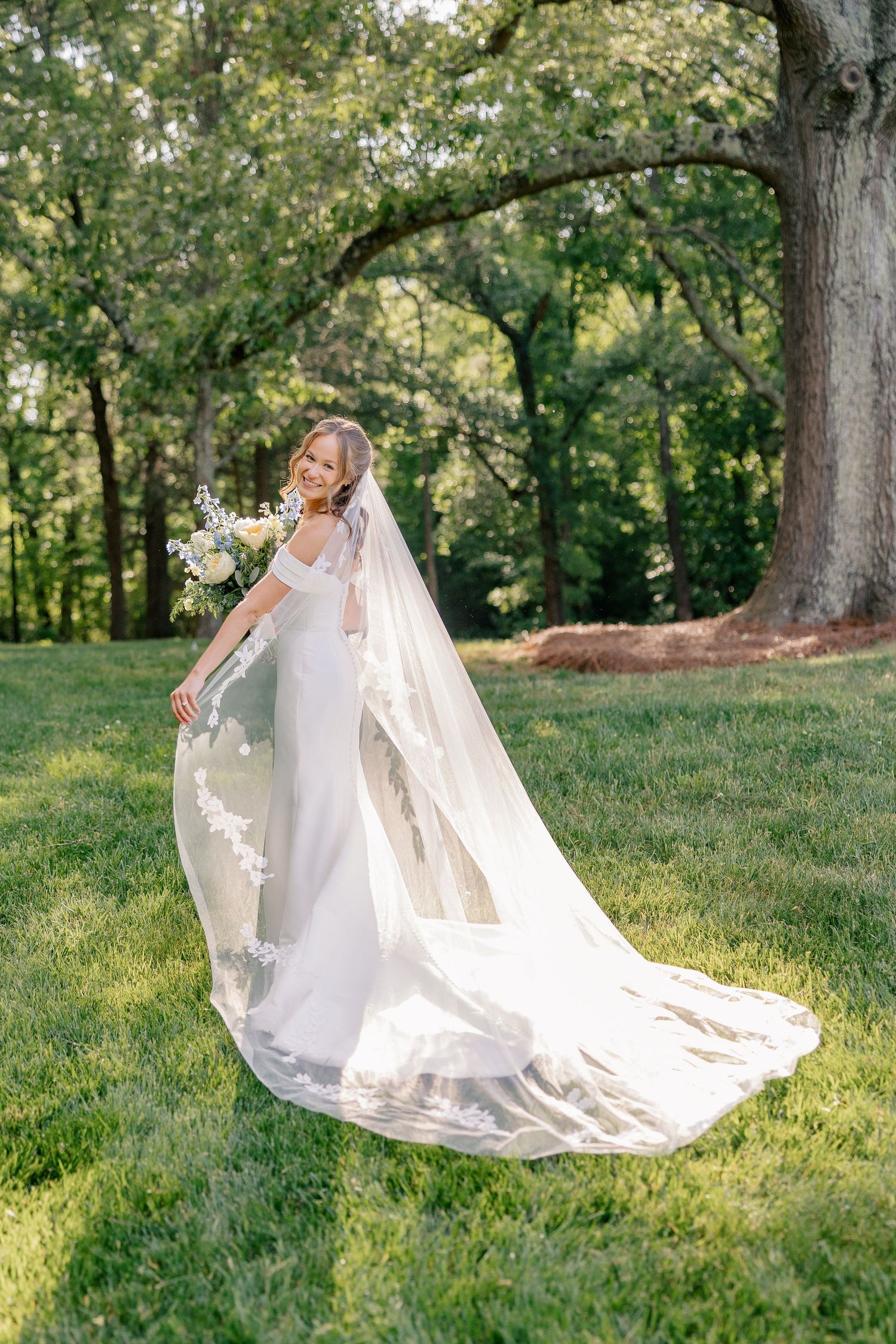 Bride in white off-shoulder gown with long lace veil holds bouquet in sunlit garden setting.