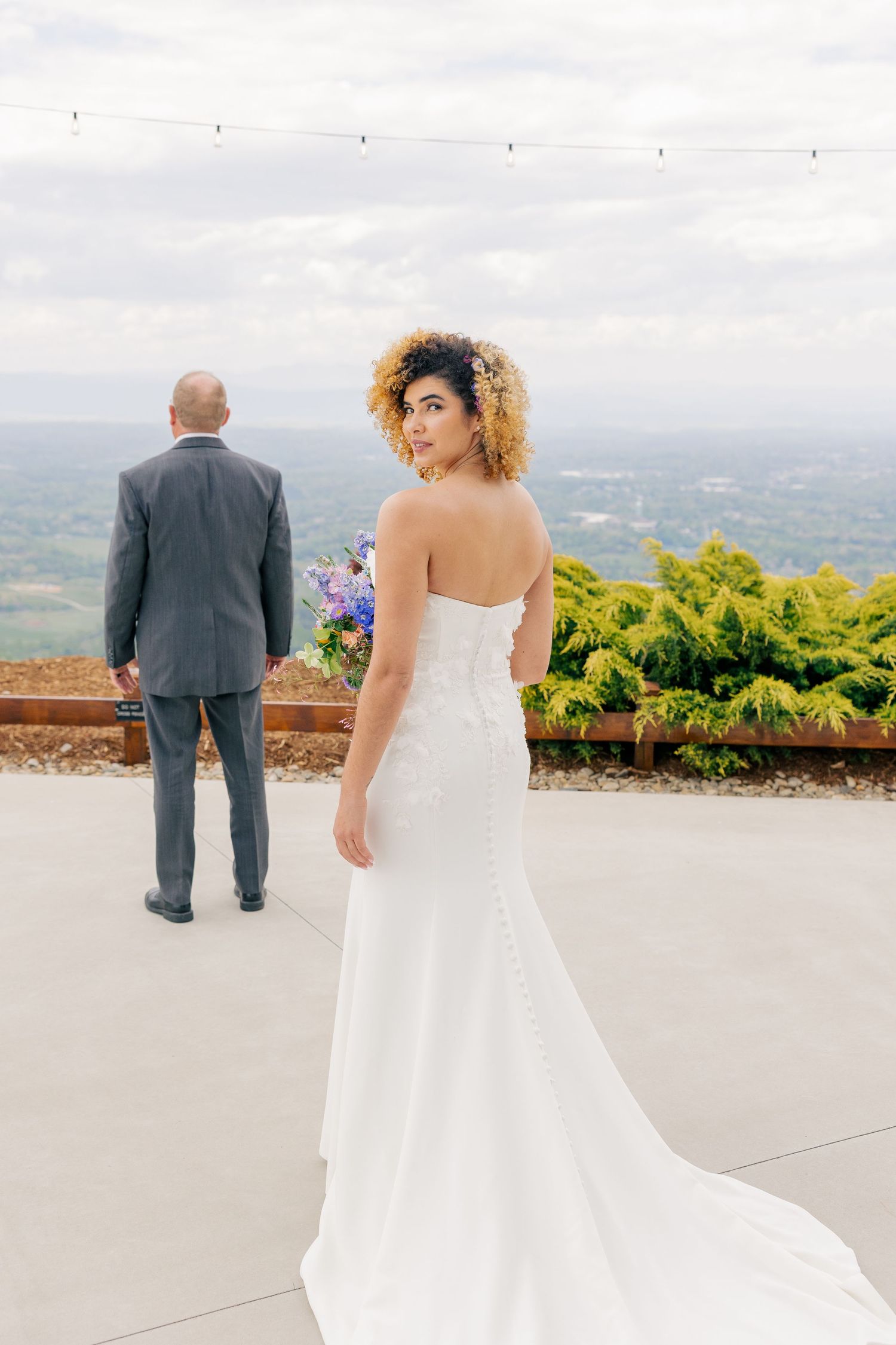 Bride in strapless white gown looks back at camera while groom in gray suit faces scenic overlook view.