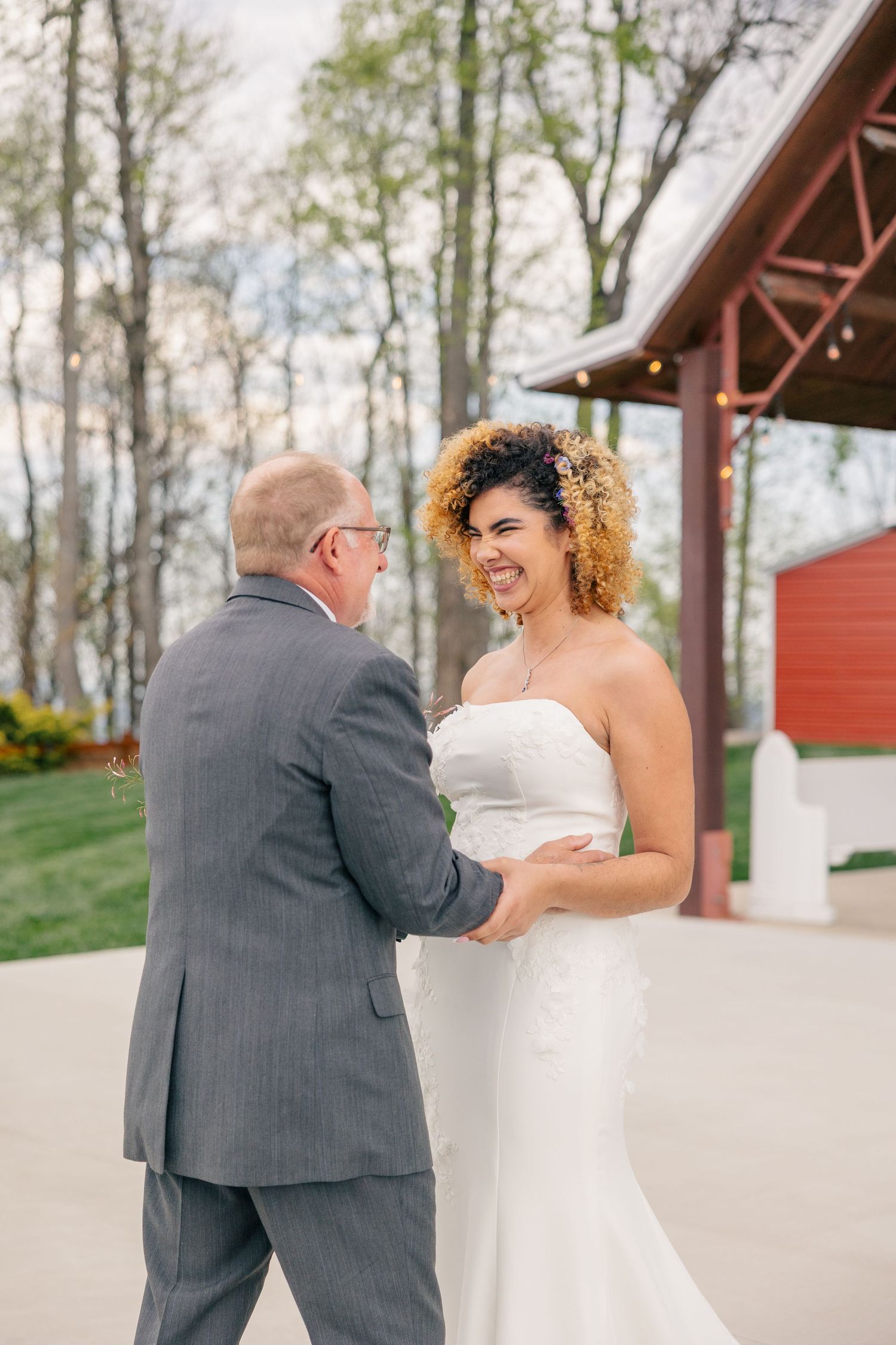 Bride in strapless white gown smiles joyfully at man in gray suit during first look at rustic outdoor wedding venue.