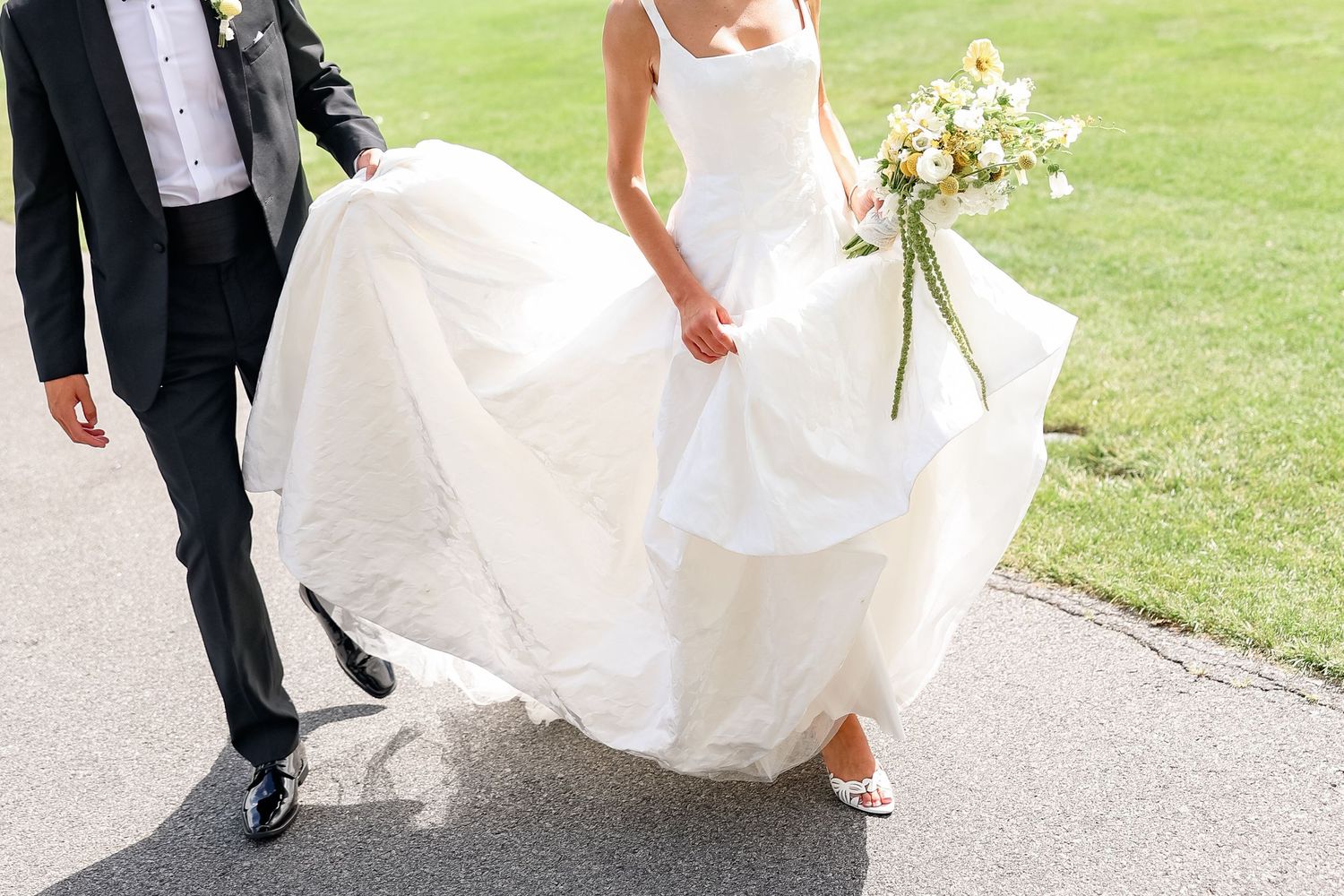 Bride in flowing white wedding gown holding sunflower bouquet walks beside groom in black suit on paved path.