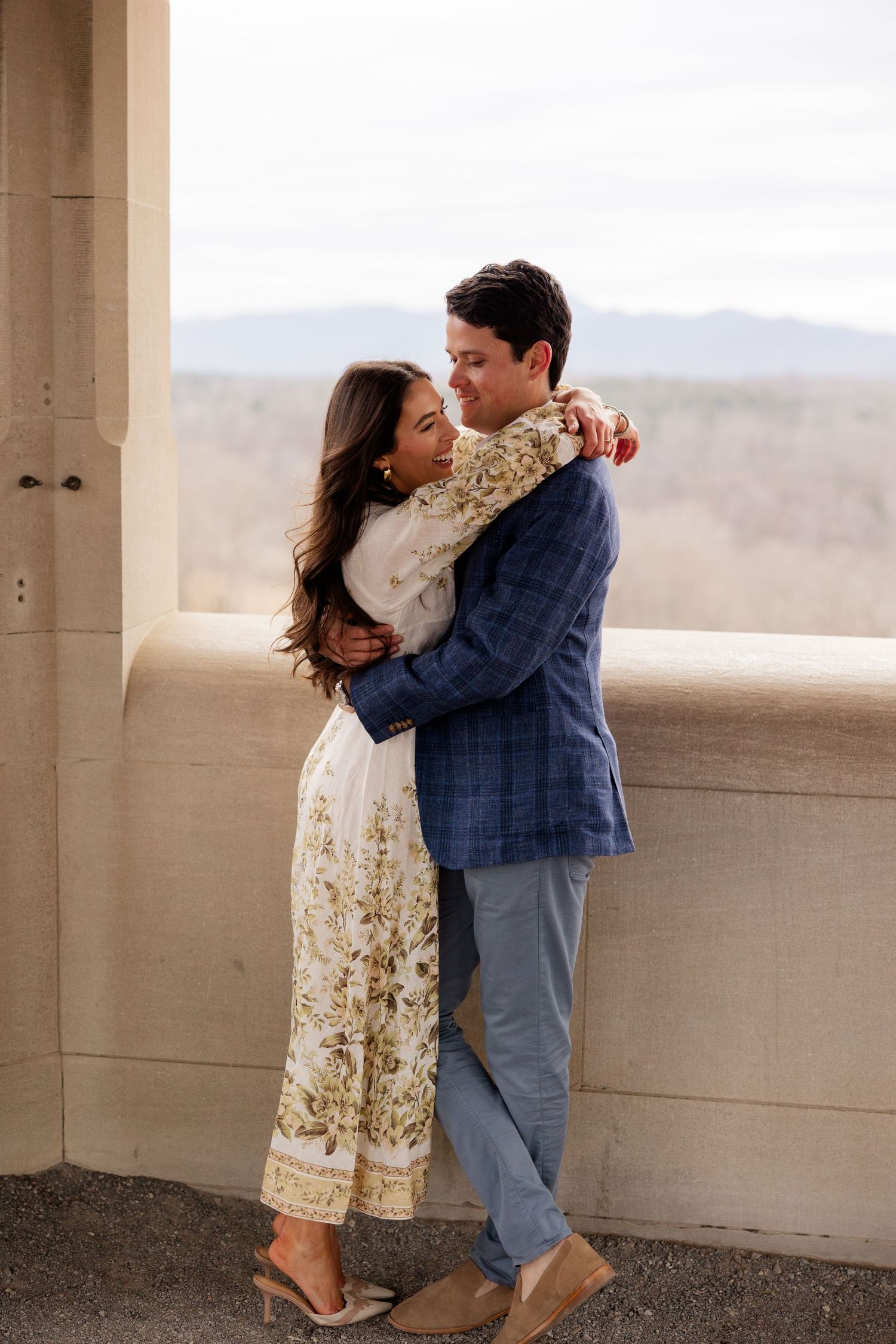 Two people embrace romantically while standing on a balcony overlooking distant mountains on a bright day.