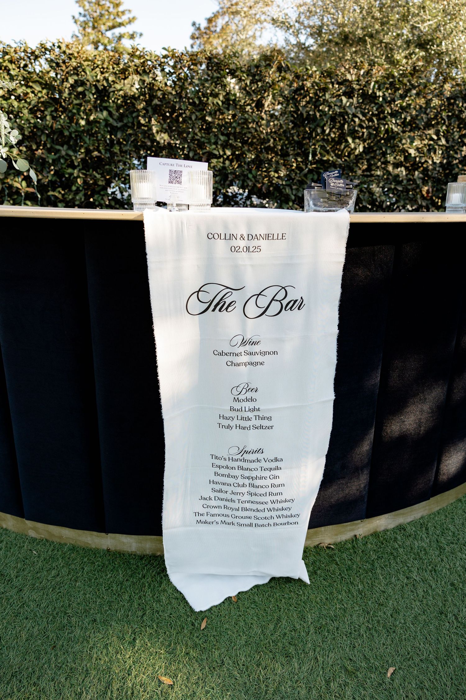 A wedding bar menu on white paper displayed against a dark backdrop with hedges in the background.