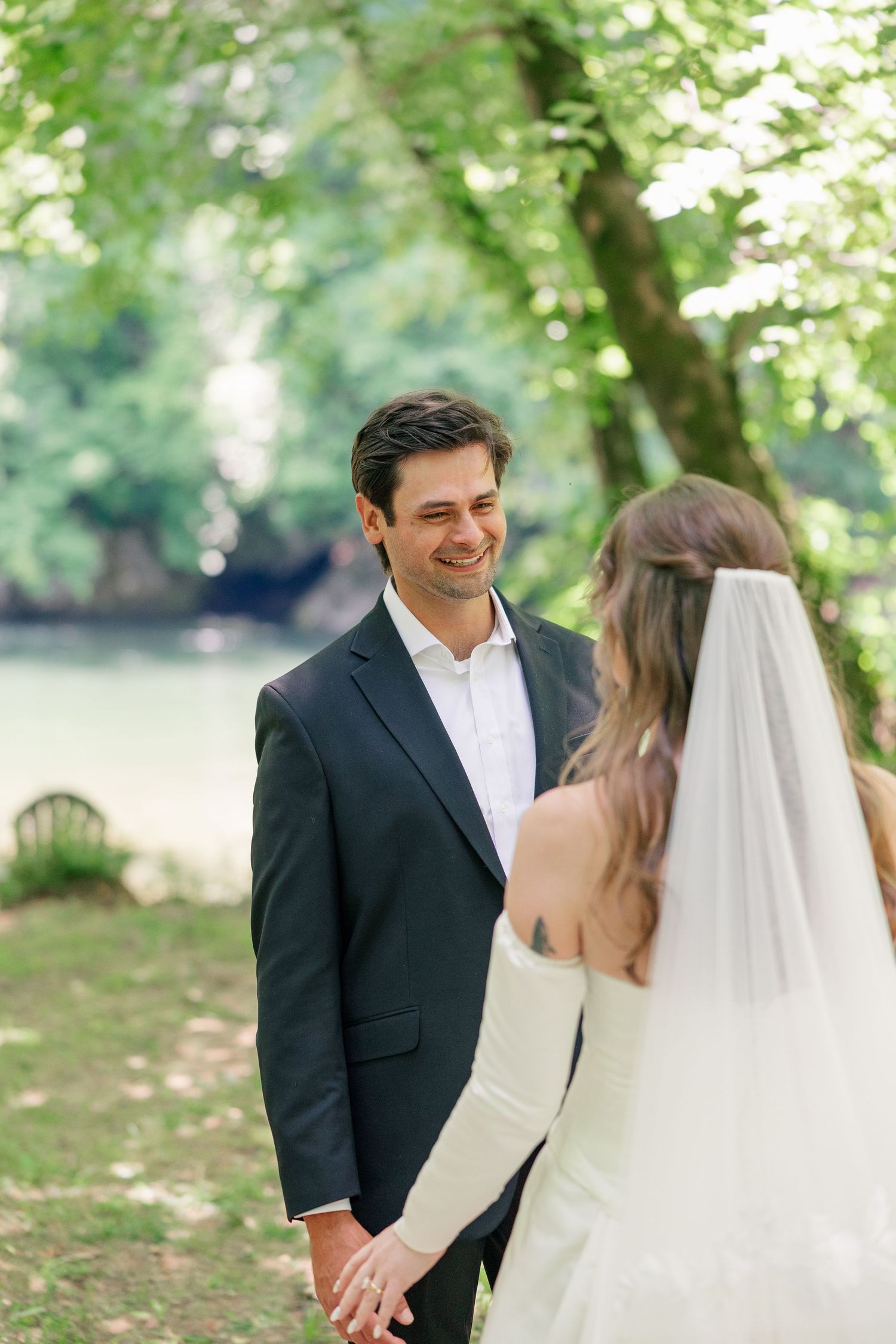 Groom in dark suit smiling at bride in white strapless gown and veil during outdoor wedding first look.