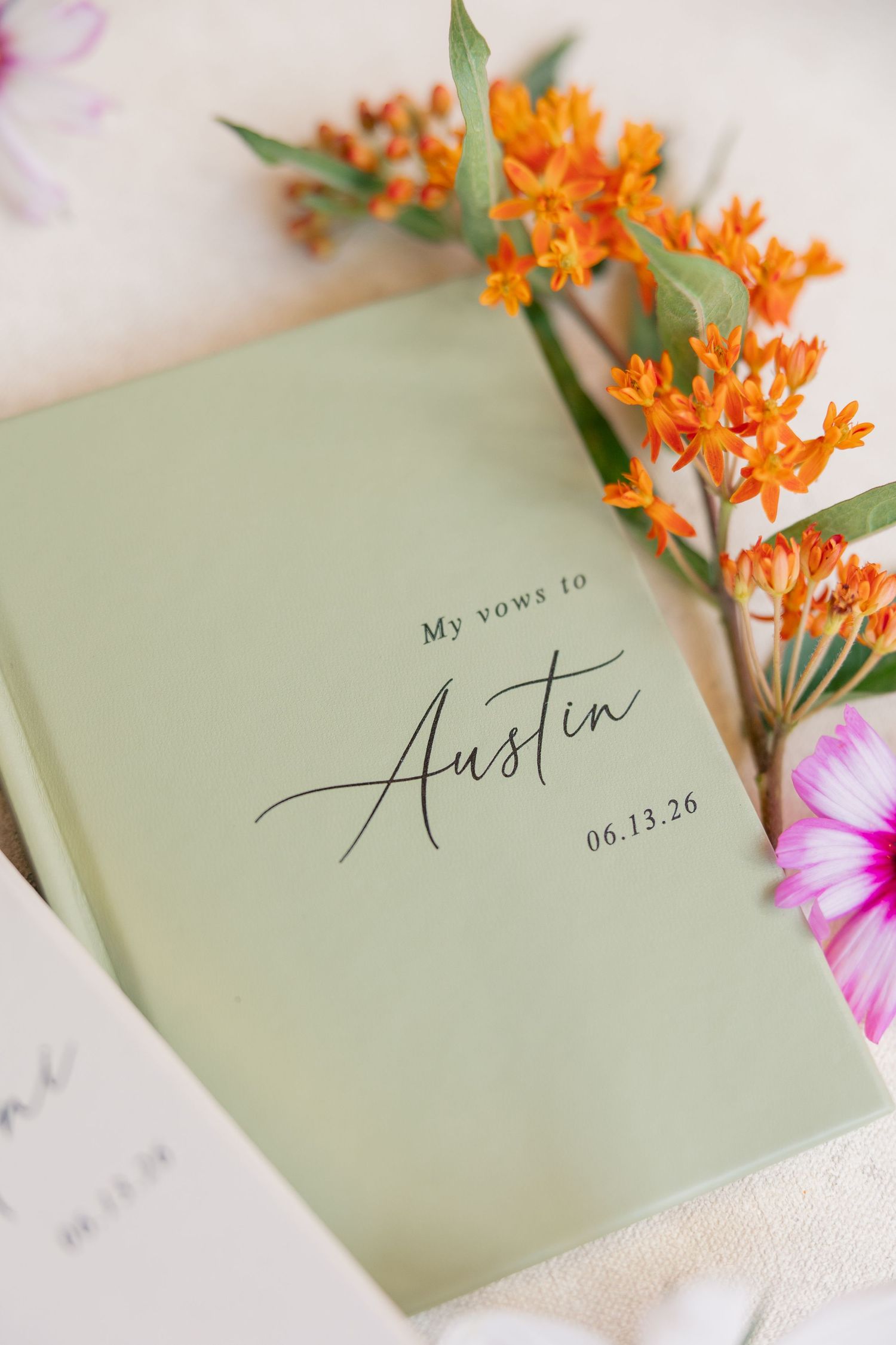 Sage green vow book personalized with Austin and wedding date 06.13.26, surrounded by orange wildflowers.