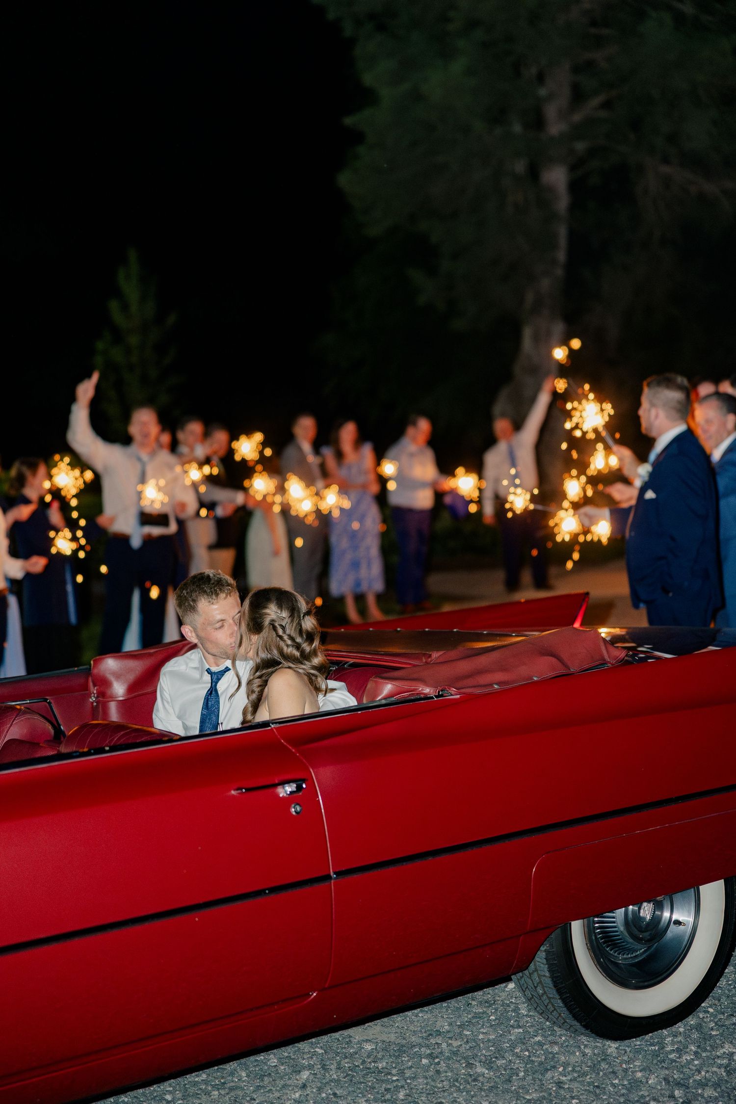 Newlyweds kissing in a classic red convertible during sparkler sendoff with cheering wedding guests at night.