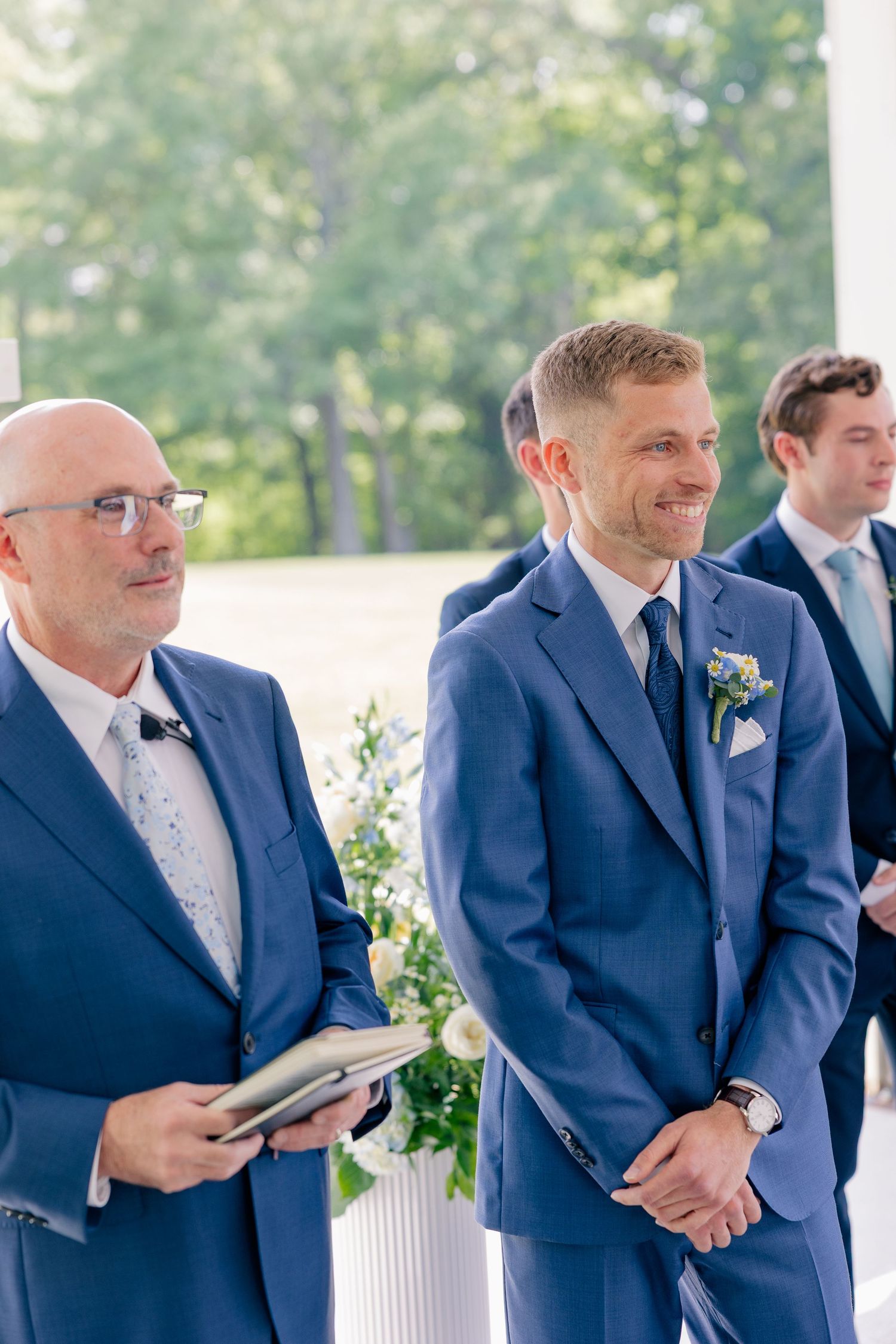 Smiling groom in blue suit standing at the altar with officiant during outdoor wedding ceremony.