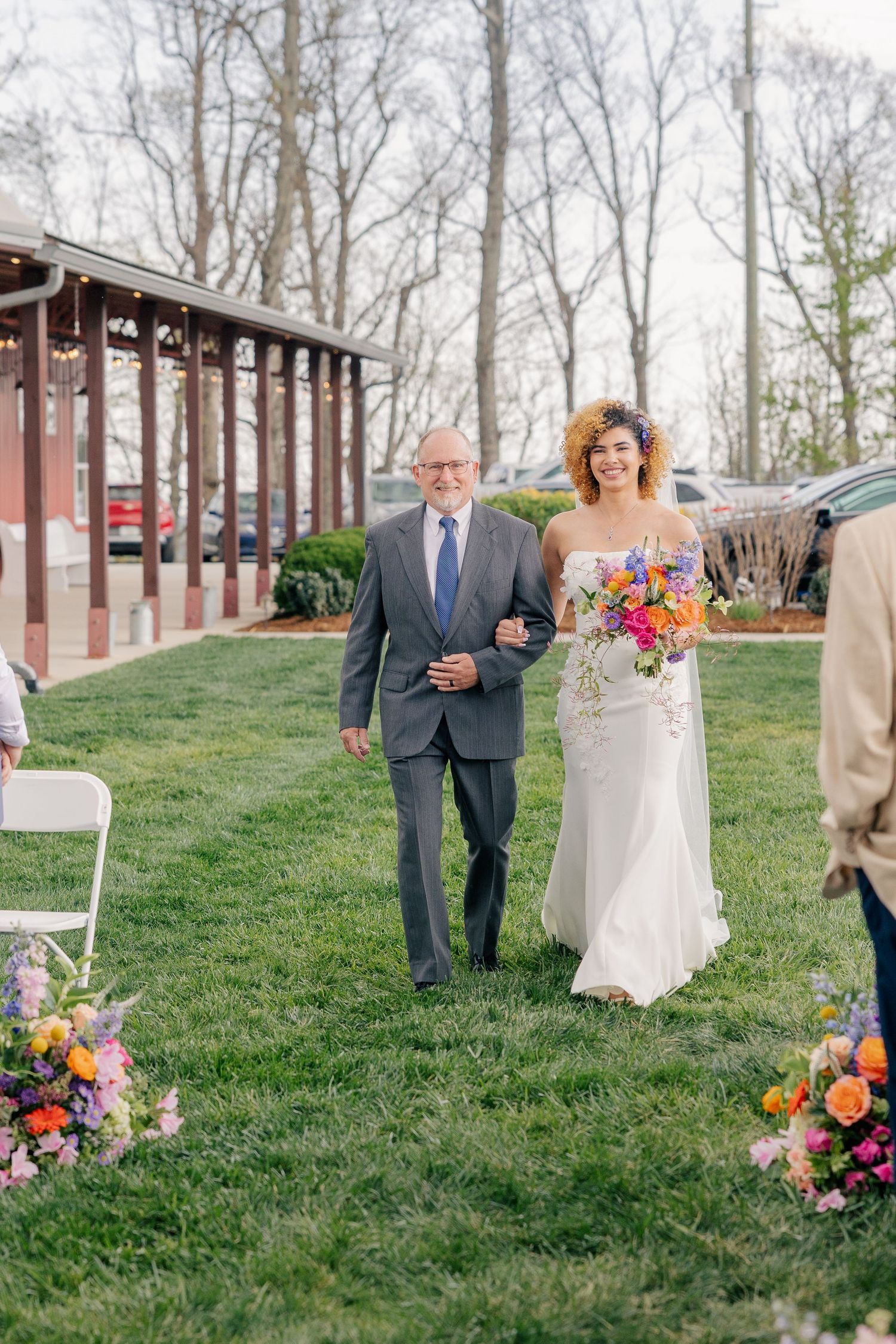 Bride walking down grass aisle escorted by father at outdoor spring wedding ceremony.