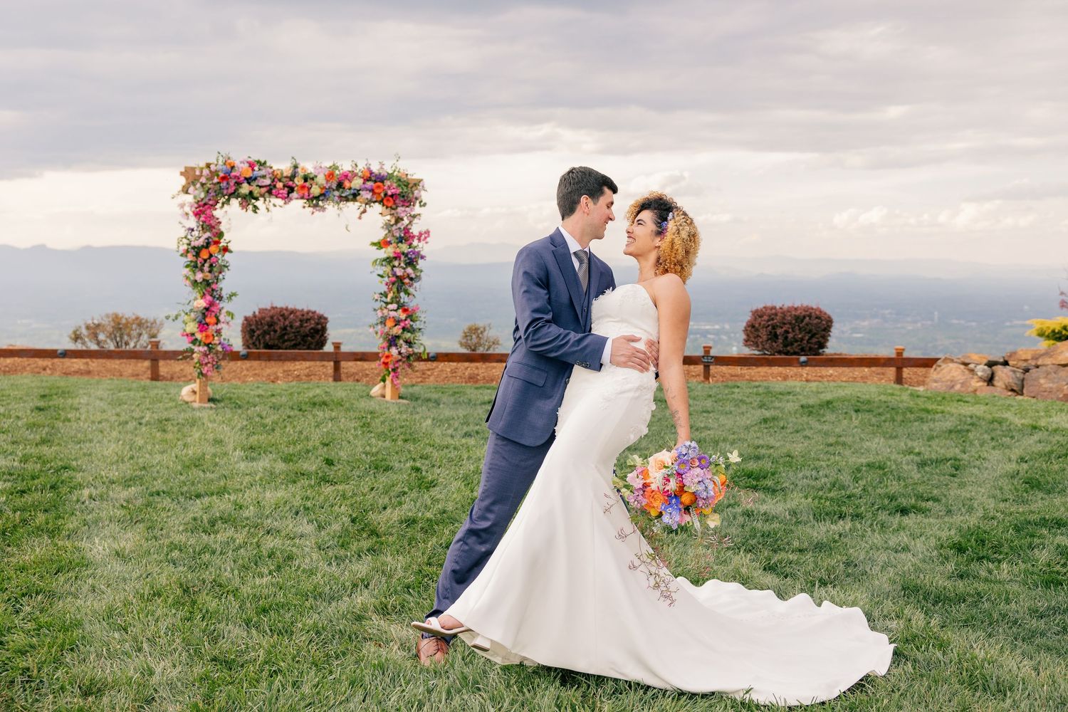 Bride and groom embracing in front of a colorful floral arch on a green lawn with mountain backdrop.