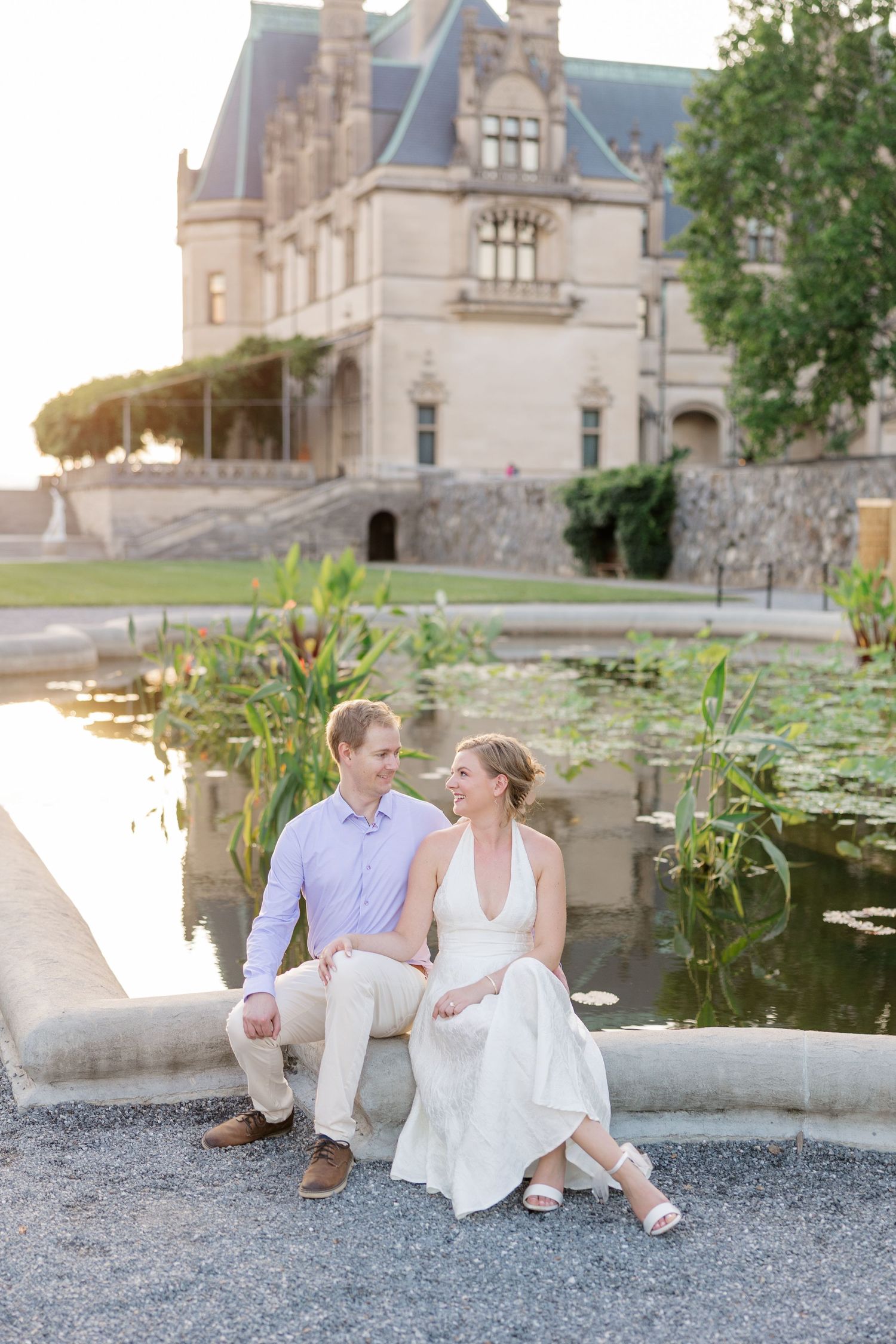 Couple sits together on stone wall by lily pond at elegant Biltmore Estate during romantic summer photo session.