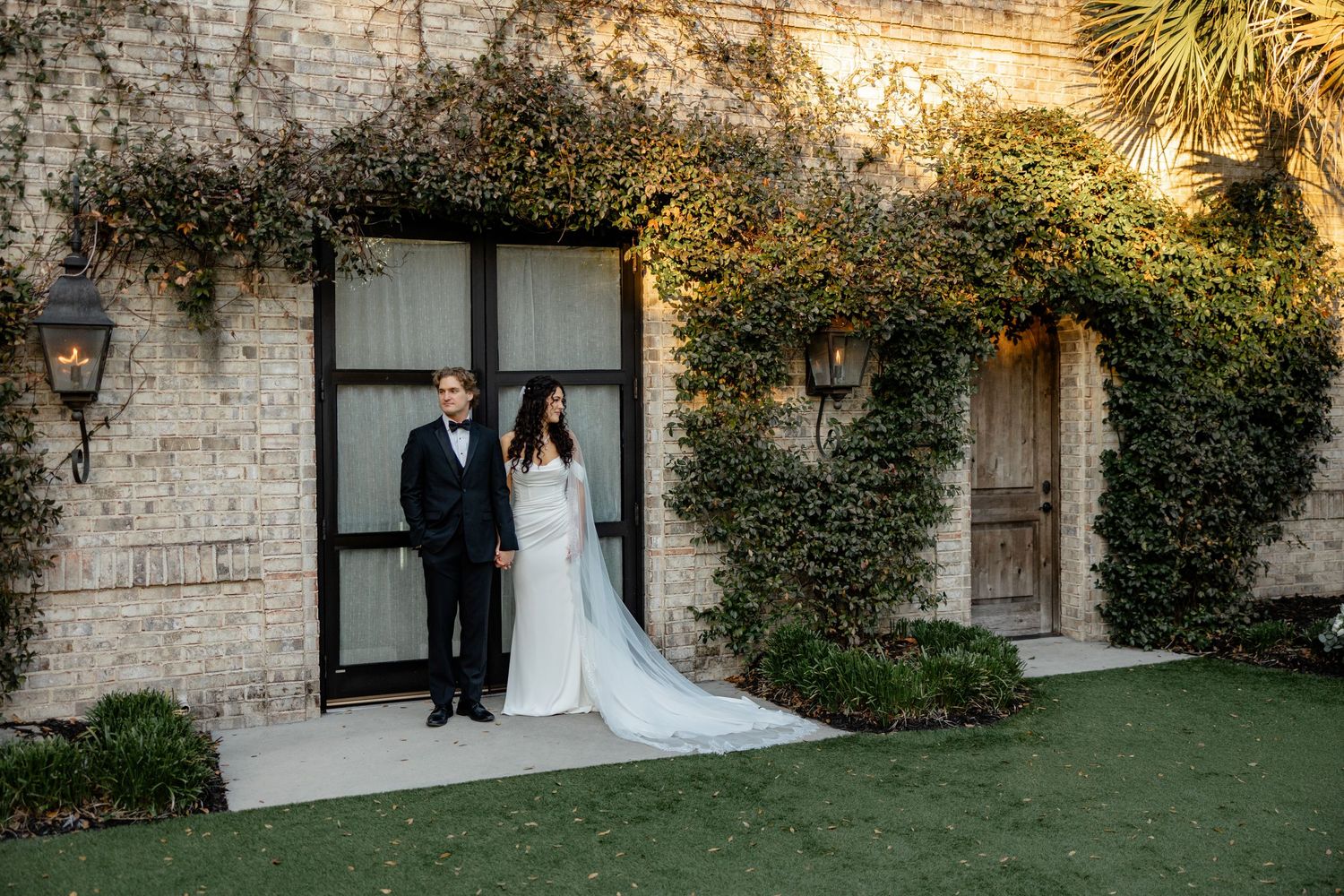 Elegant couple poses in front of an ivy-covered stone building at dusk.
