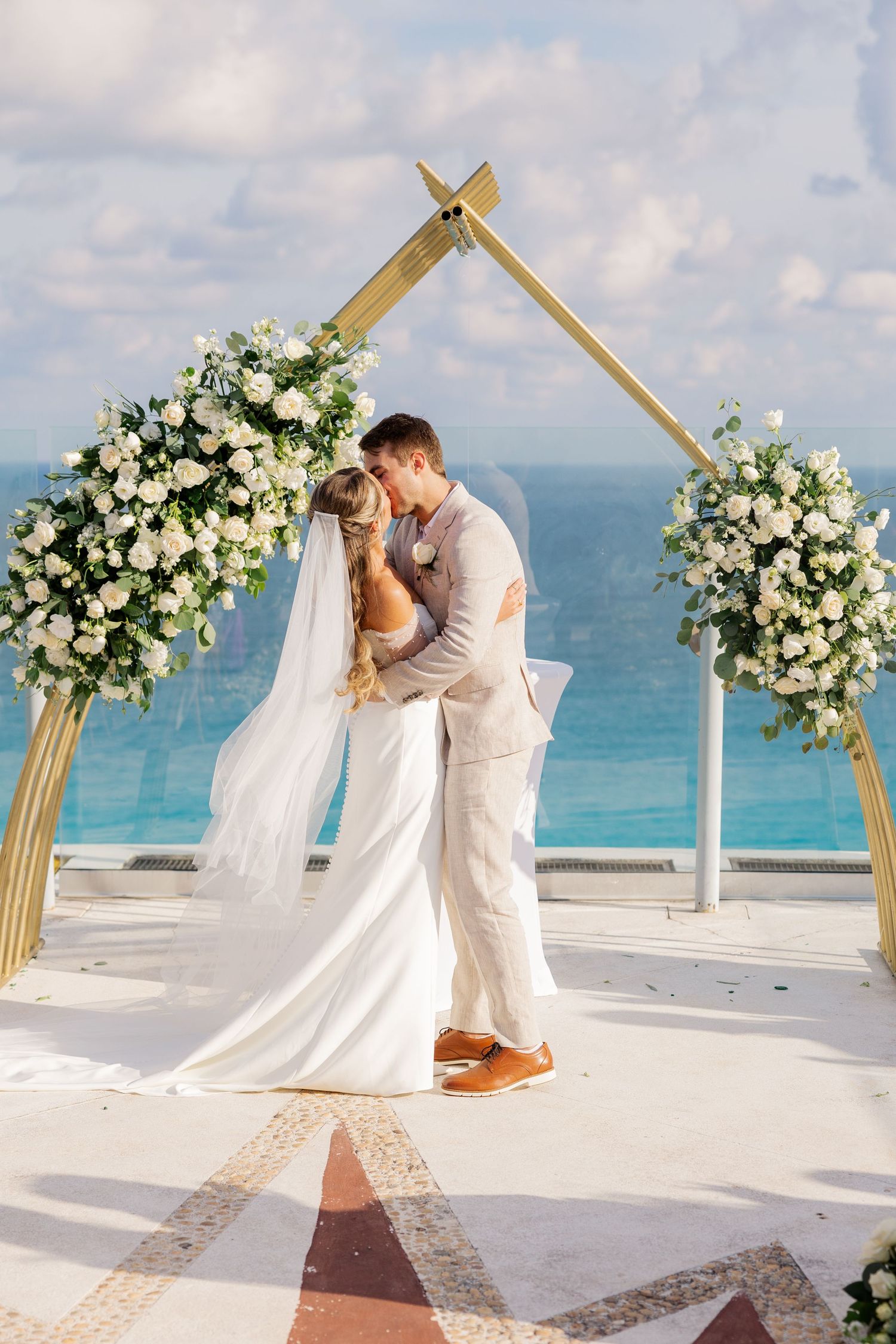 Bride and groom share first kiss under gold floral arch with white roses, ocean backdrop at outdoor beach wedding ceremony.
