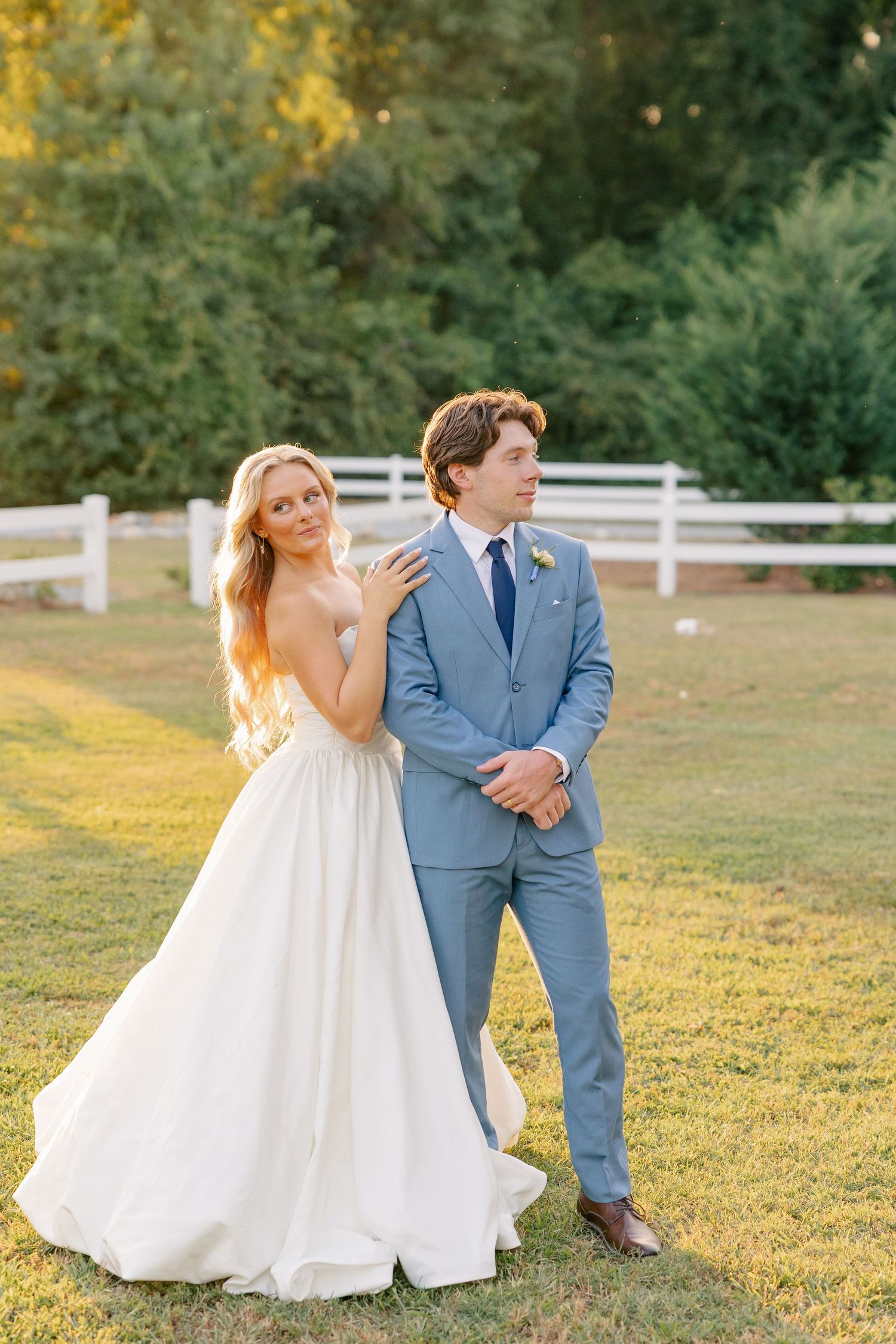 Bride in strapless white gown and groom in light blue suit pose together on a sunlit green lawn at golden hour.