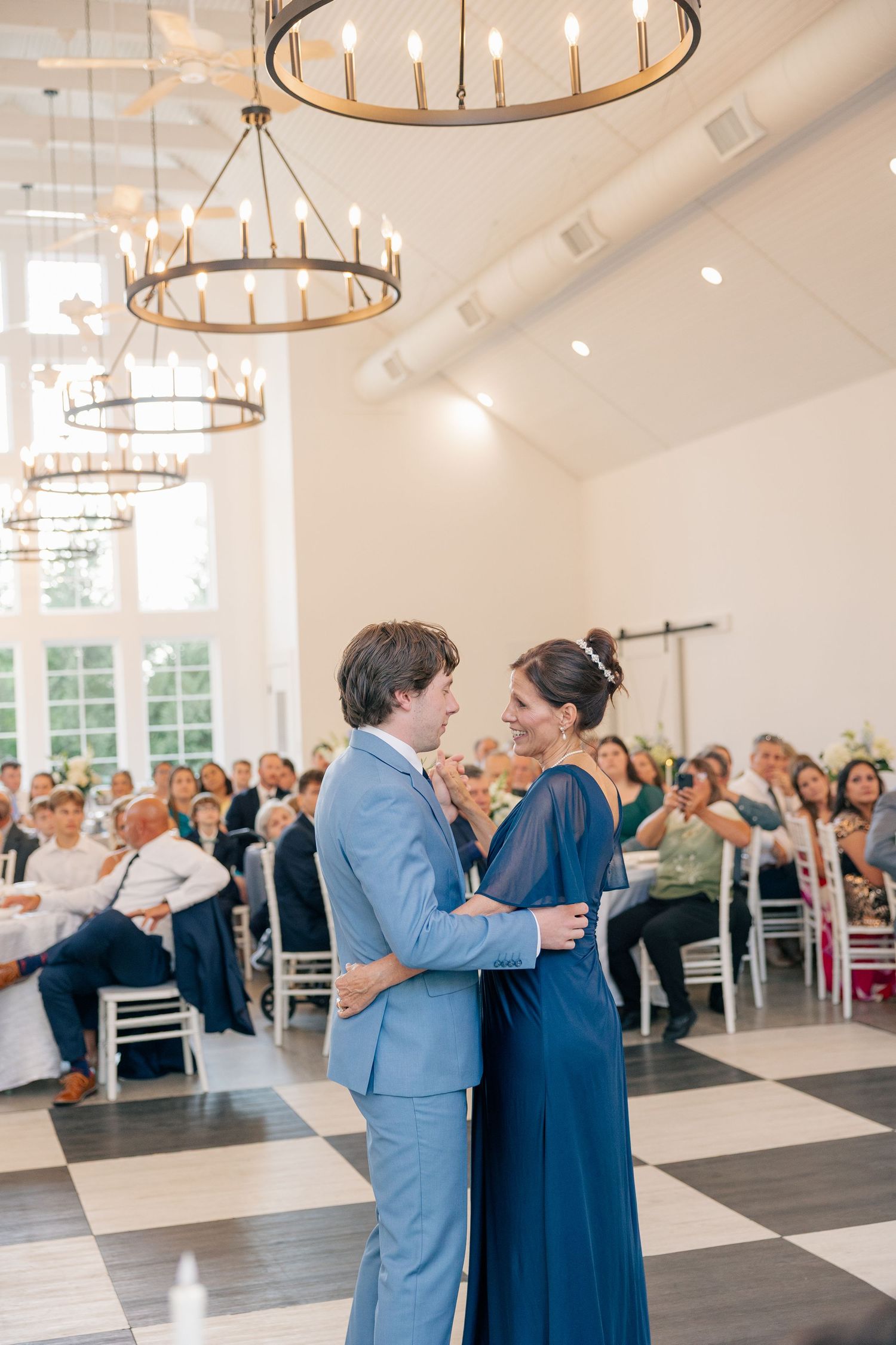 Groom in light blue suit shares tender mother-son dance on checkered ballroom floor.