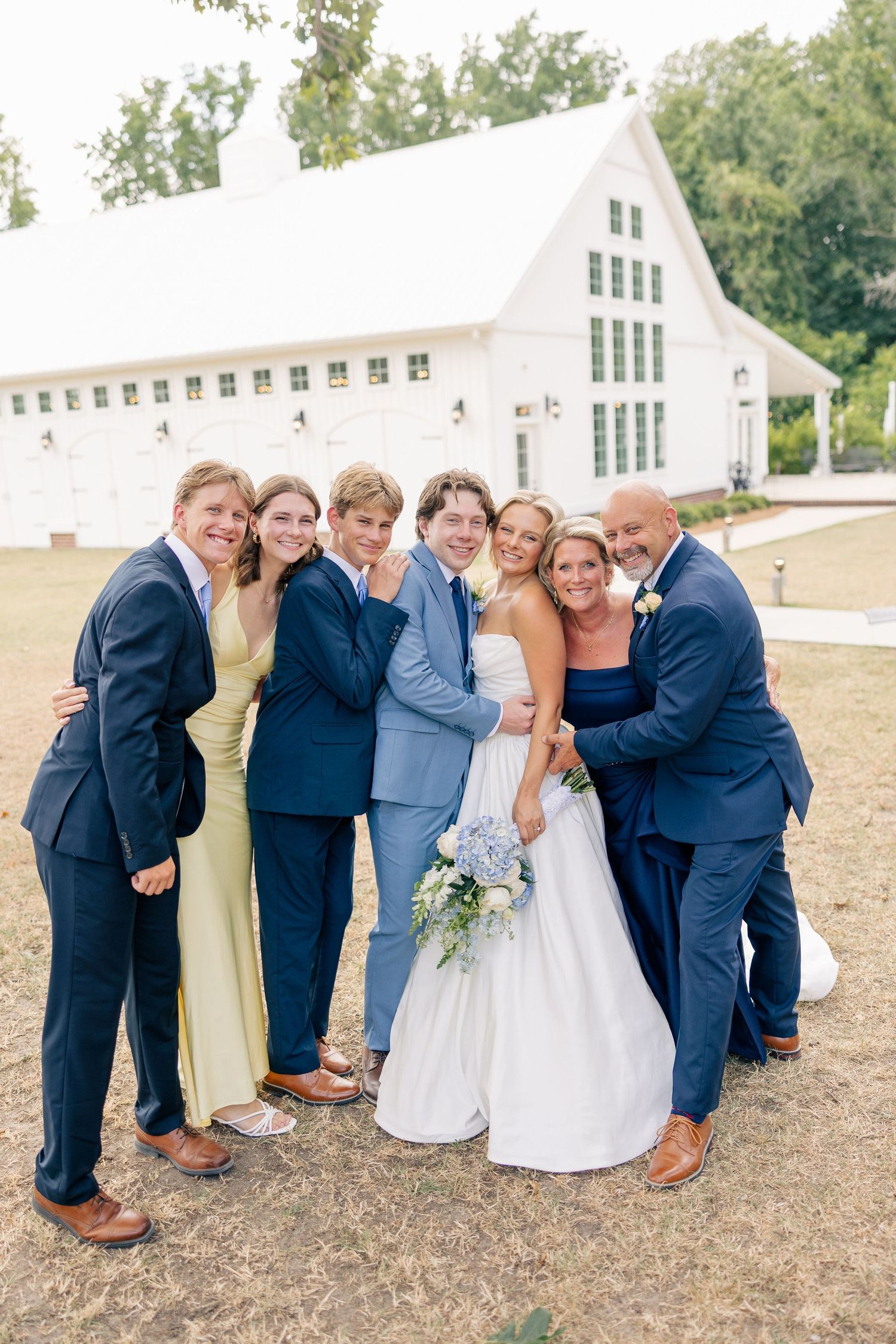 Newlyweds celebrate with family group photo outdoors in front of white barn wedding venue.