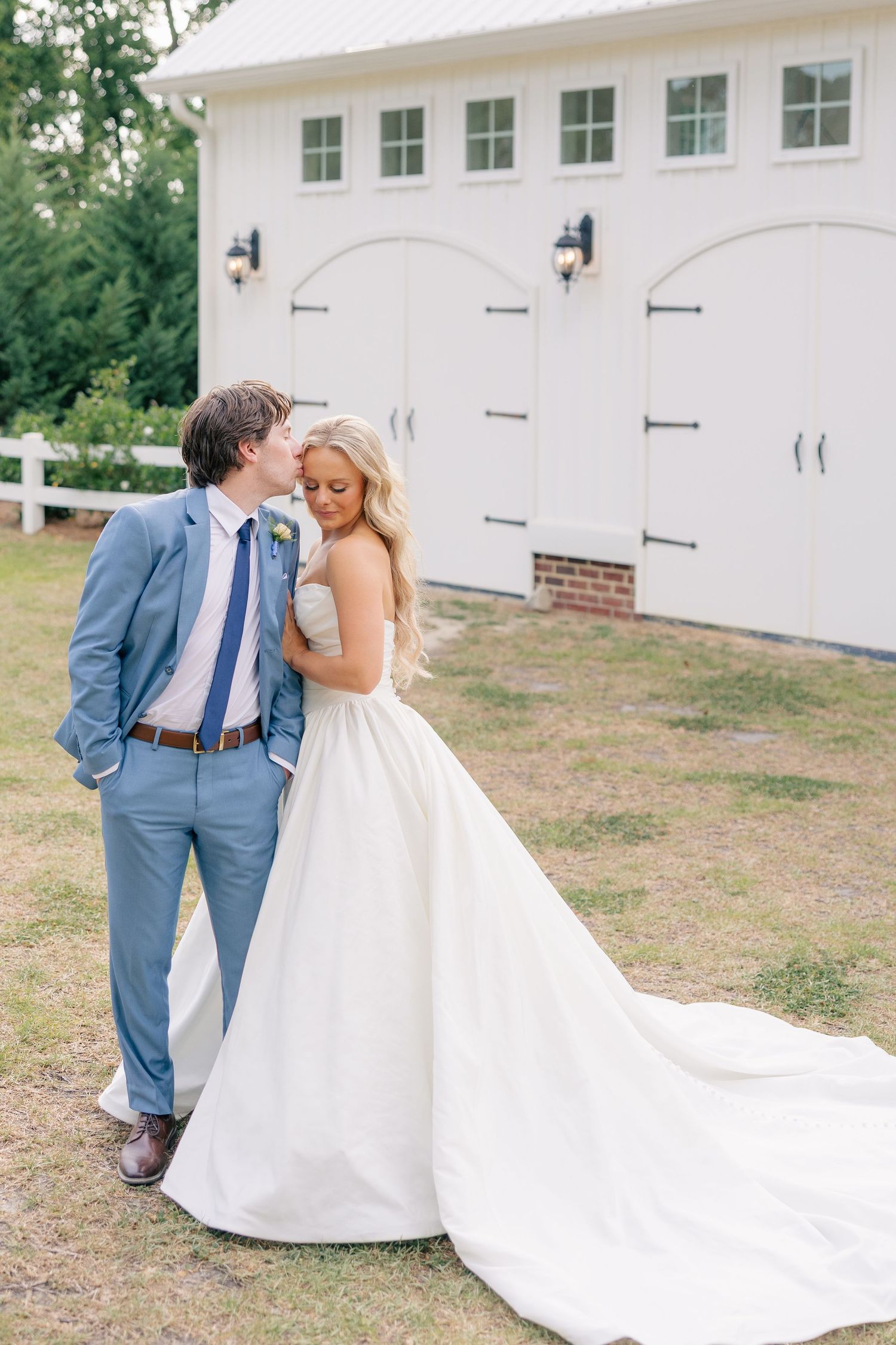 Groom in light blue suit kisses bride in strapless ball gown outside white barn venue on wedding day.