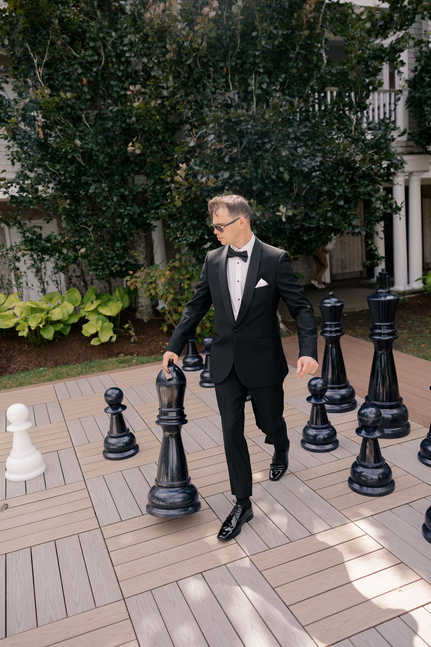Groom in black tuxedo posing with oversized black chess pieces on outdoor wooden deck.