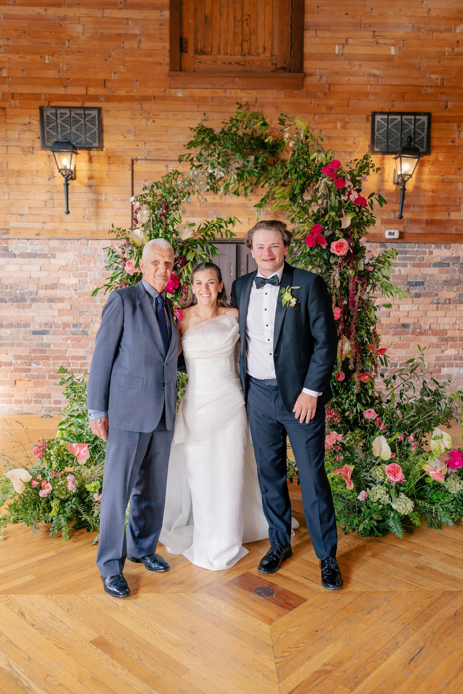 Bride, groom, and officiant pose before lush floral arch with pink roses in rustic venue.