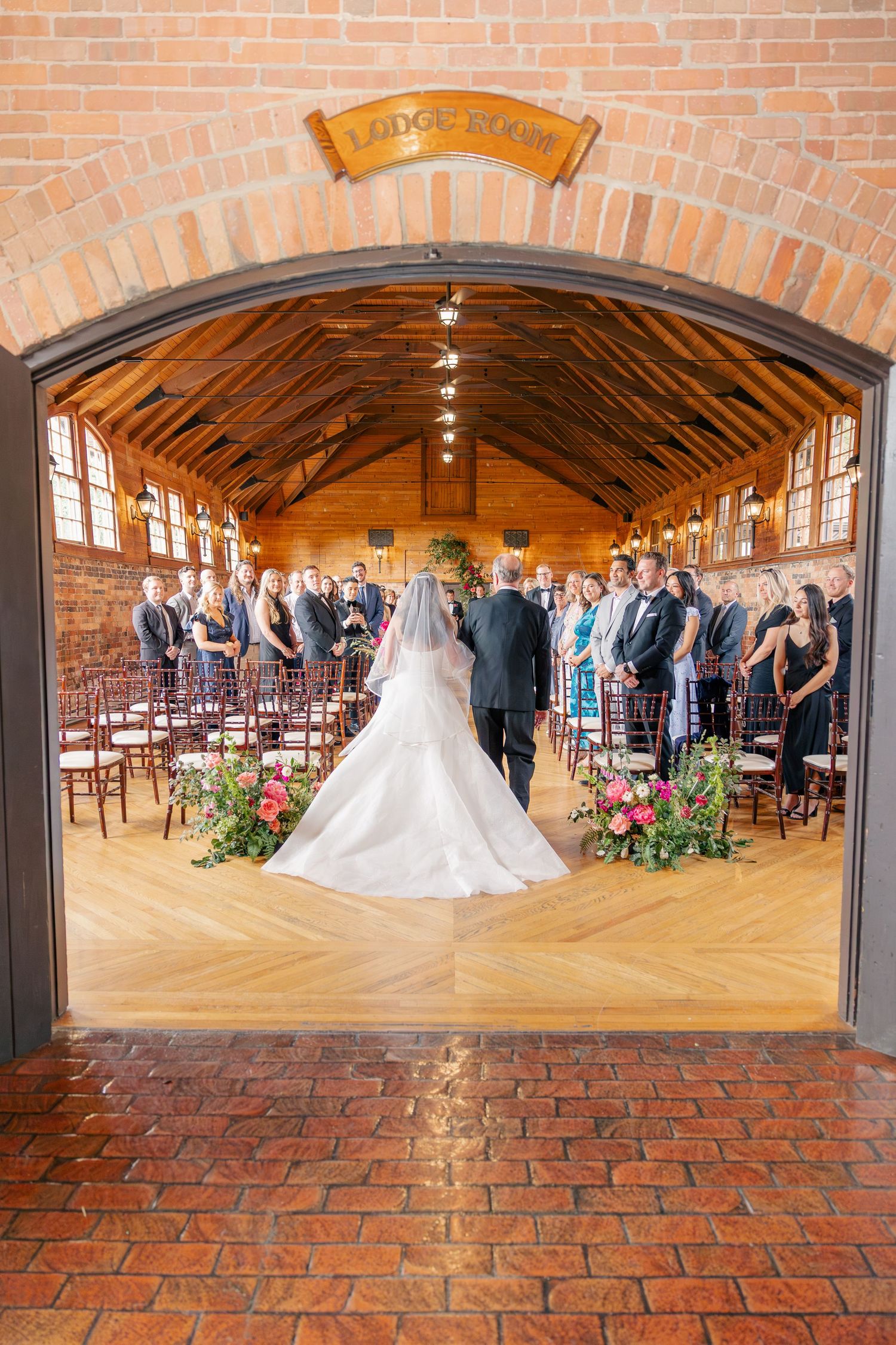 Bride walks down the aisle in elegant white gown at indoor barn wedding venue with wooden floors.