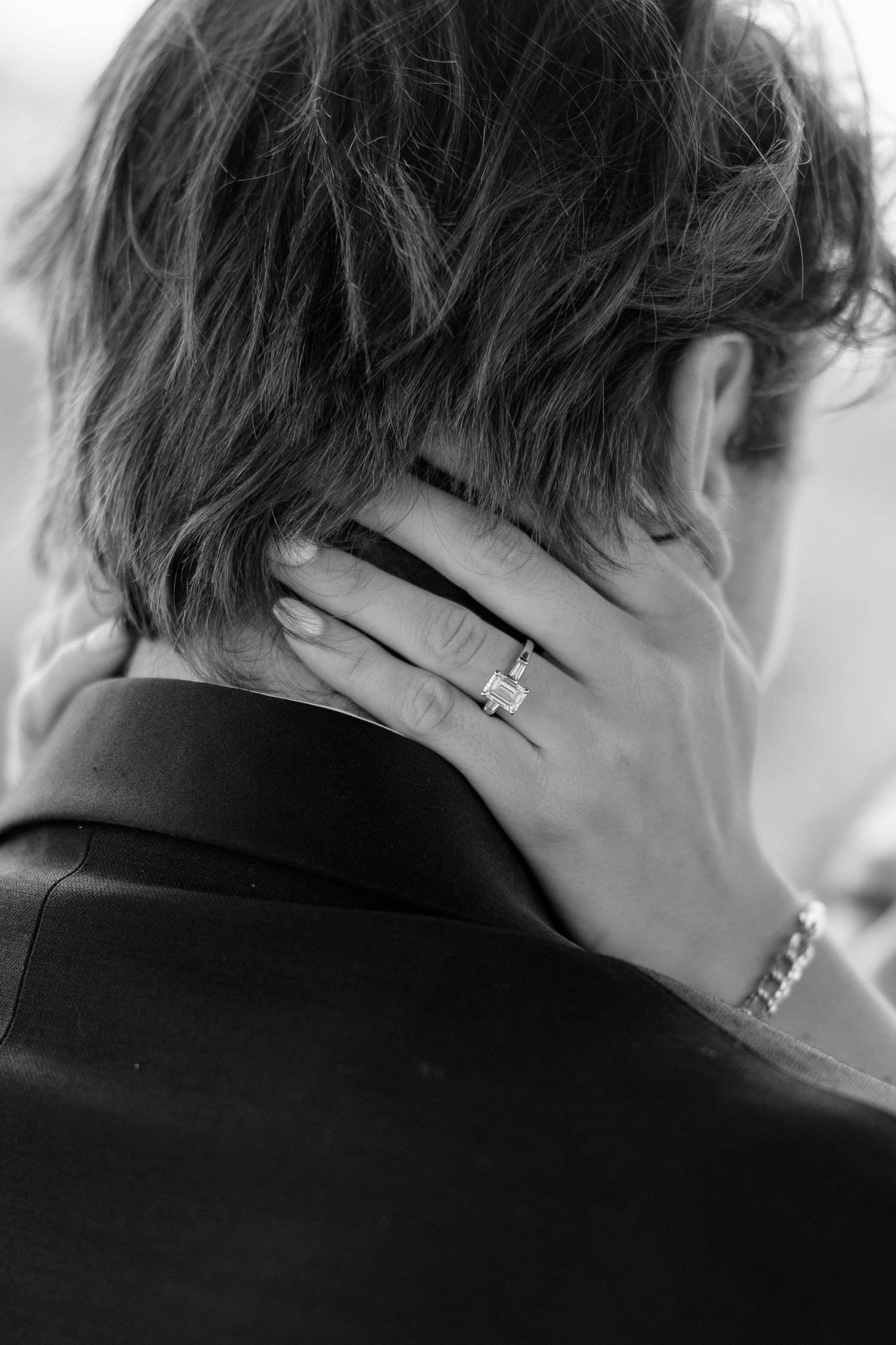Black and white close-up of brides hand with emerald cut engagement ring resting on grooms neck.