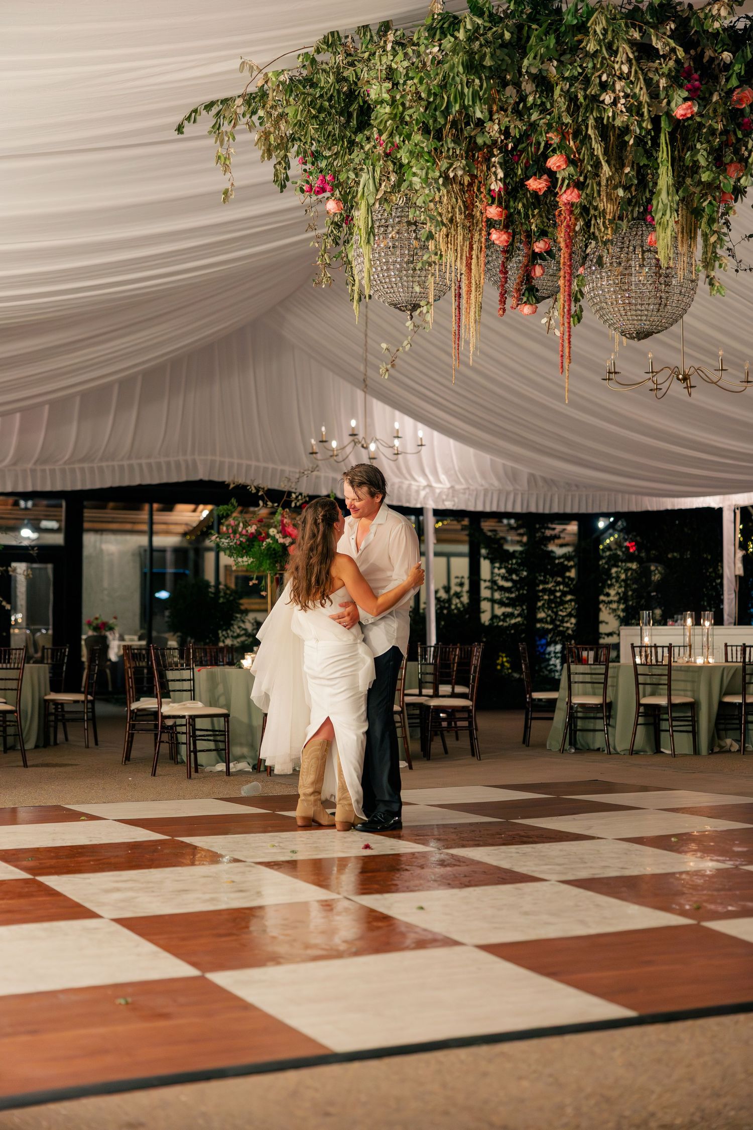 Bride and groom share their first dance on a checkered floor under floral chandelier.