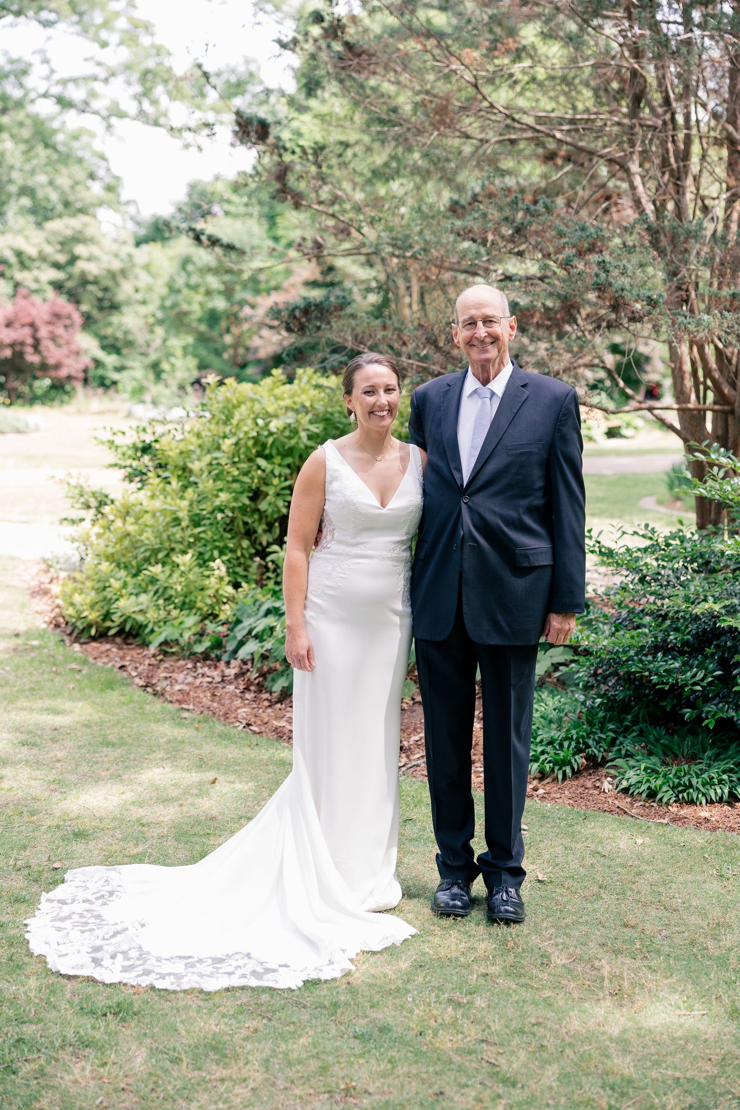 Bride in elegant white gown with lace train poses with older gentleman in navy suit outdoors.