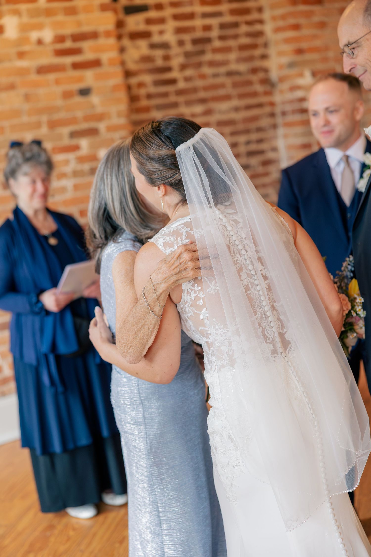 Emotional bride embraces mother in silver dress during heartfelt wedding ceremony moment.