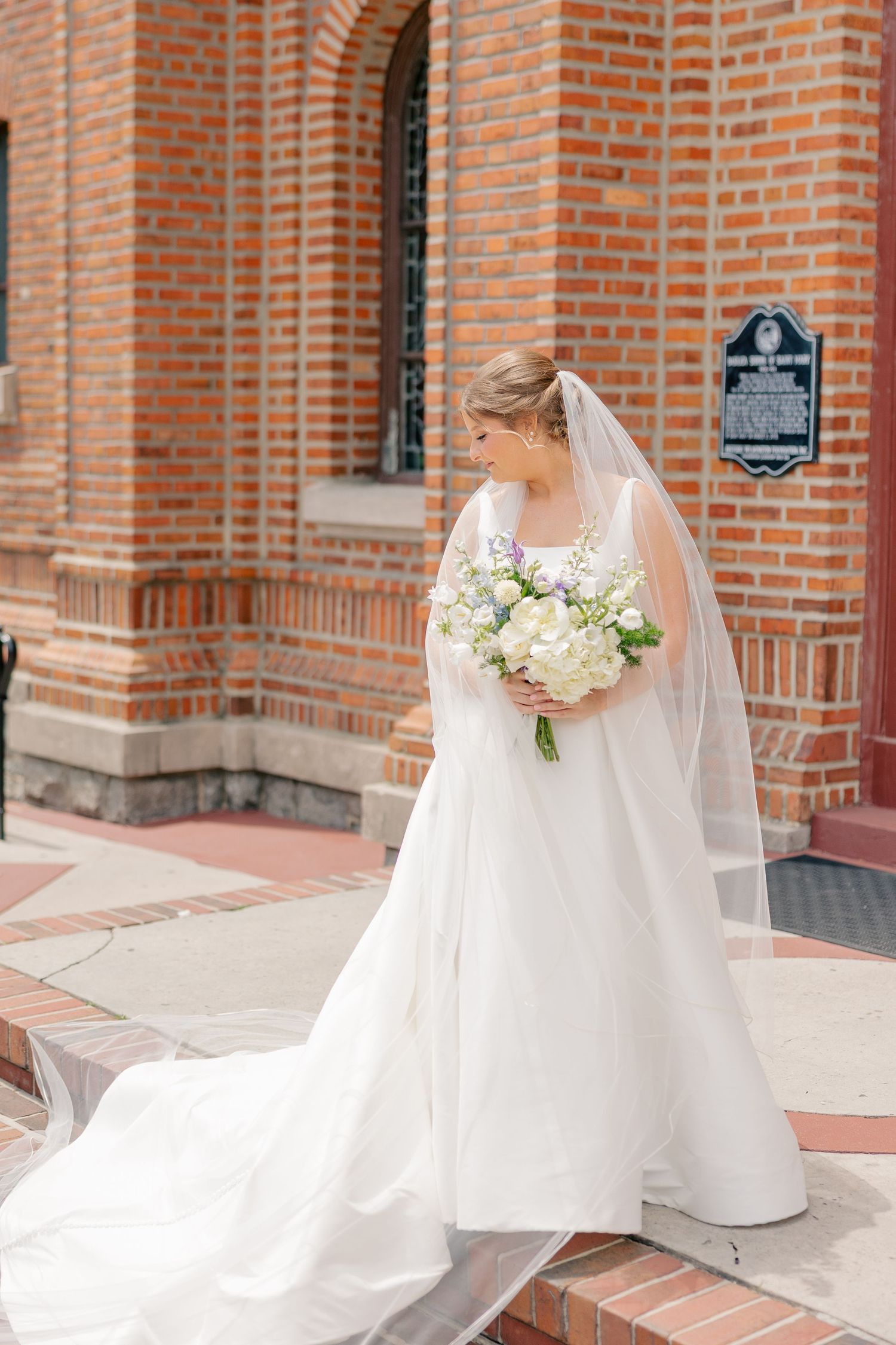 Bride in a white ballgown with long veil holds a white floral bouquet on the steps of a brick church.