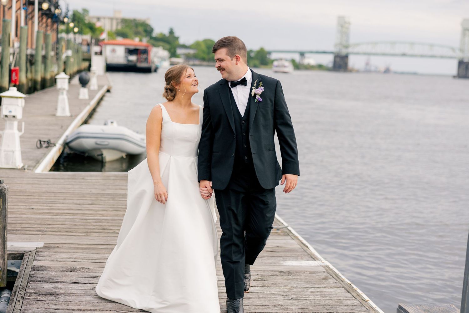 Bride and groom walk hand-in-hand on a waterfront dock, smiling at each other on their wedding day.