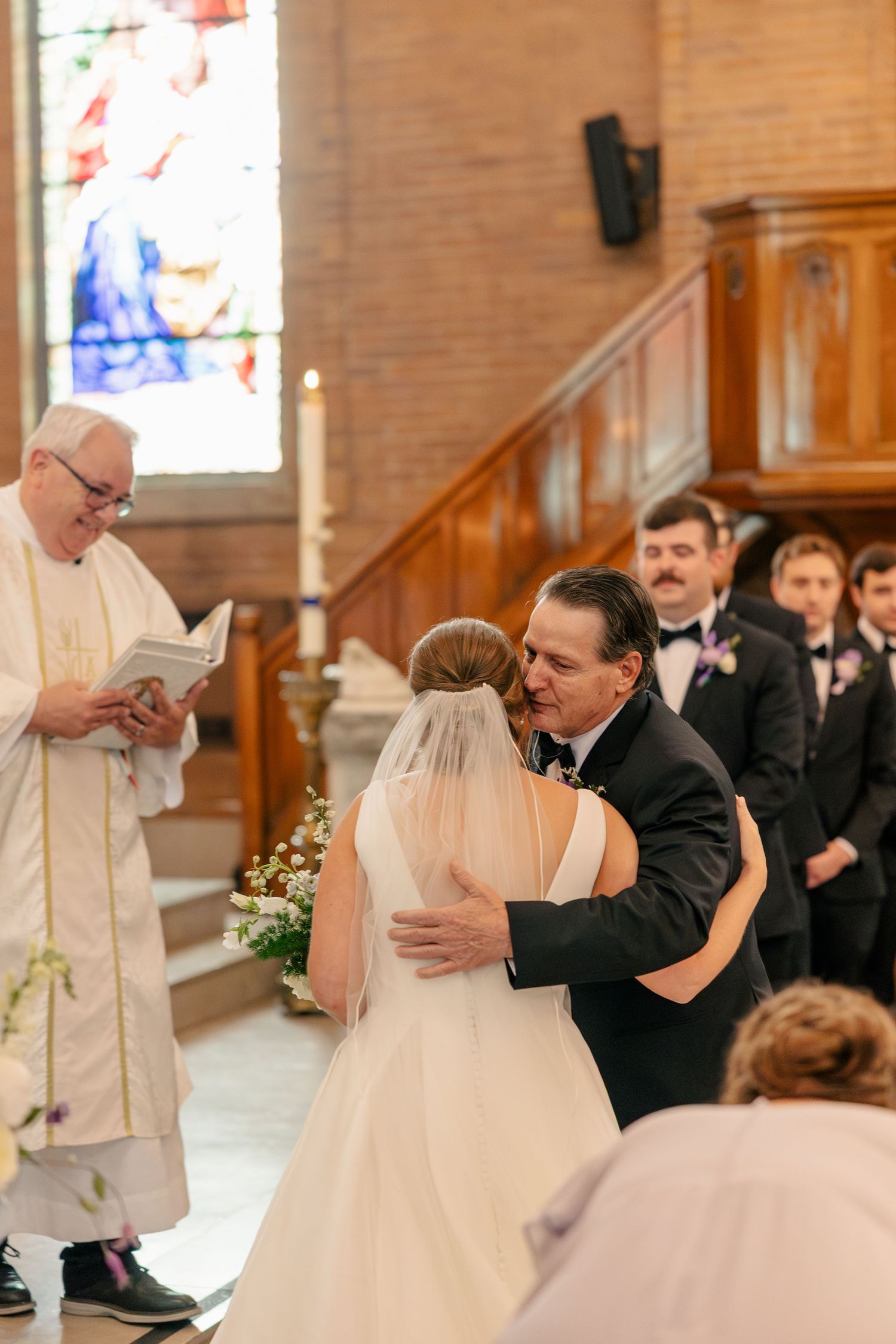 Bride in white gown and veil shares emotional hug with father at church altar during wedding ceremony.