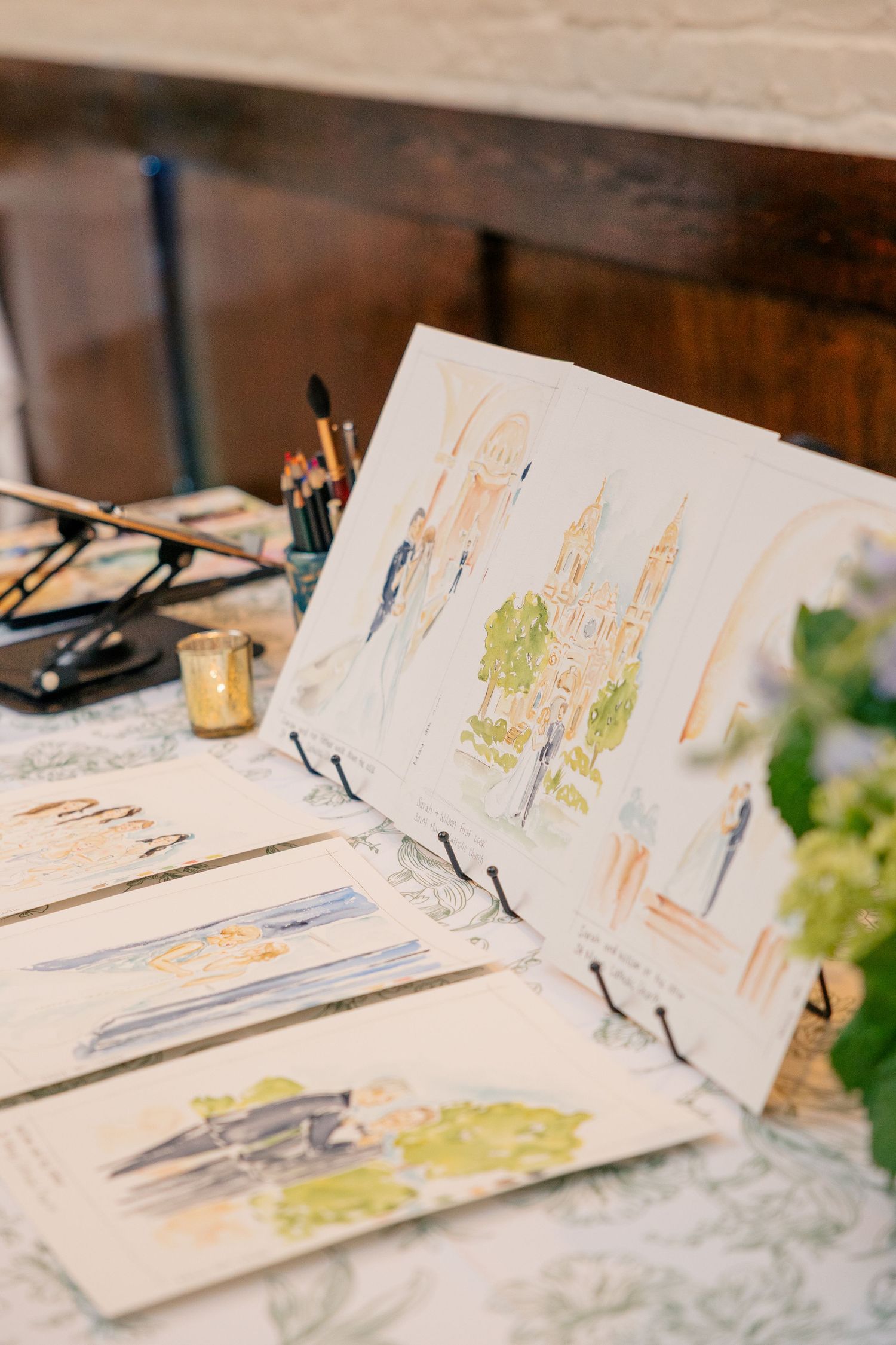 Watercolor wedding illustrations displayed on easels at reception table with paintbrushes and art supplies nearby.