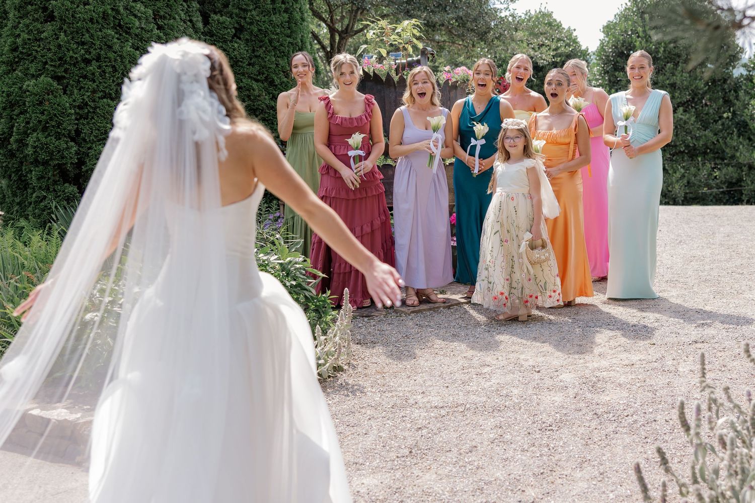 Bridesmaids in colorful dresses stand together watching bride in white gown.