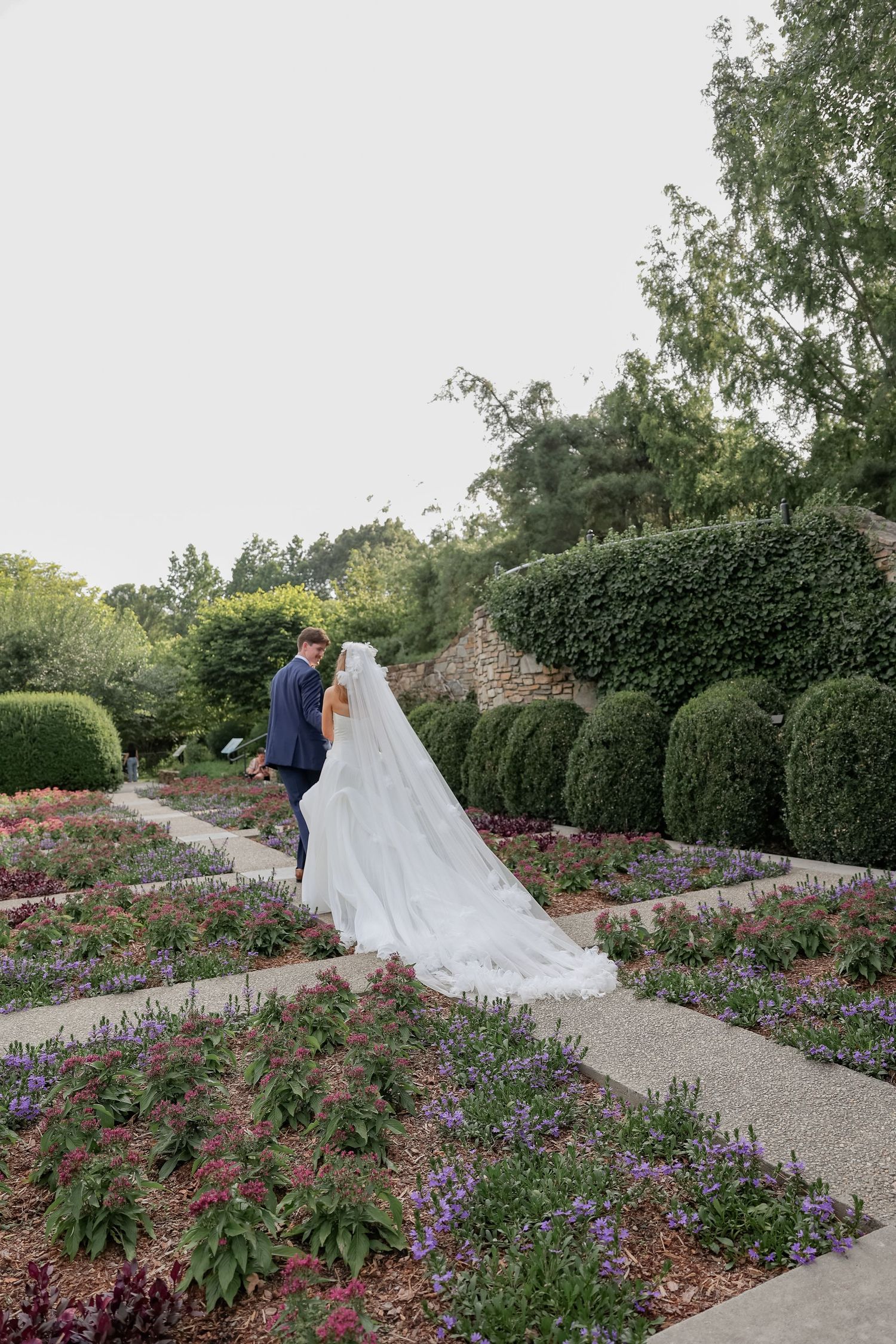 Wedding couple walking through a manicured garden path lined with colorful flowers.