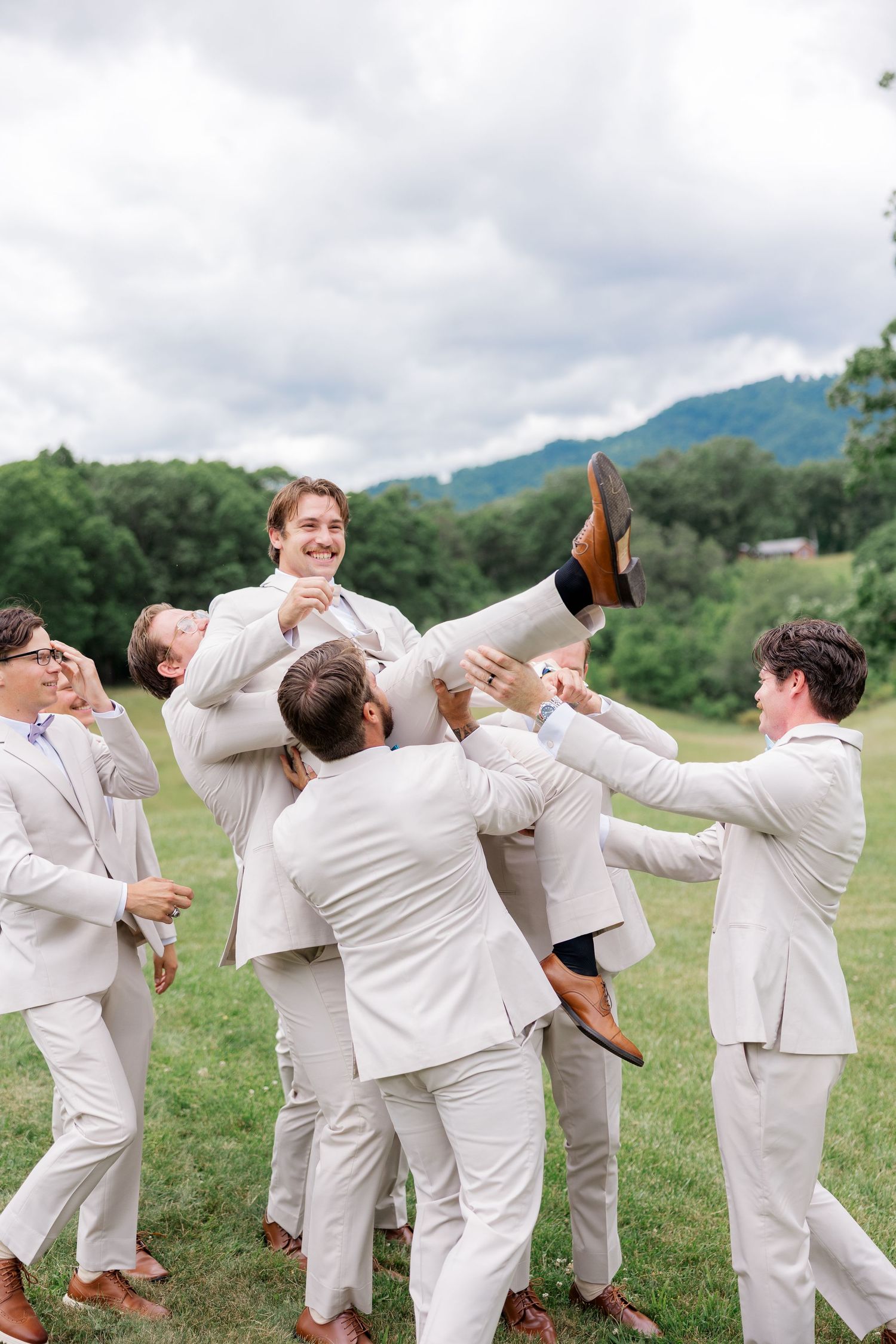 Groomsmen in light colored suits posing playfully on a grassy hillside with mountains in the background.