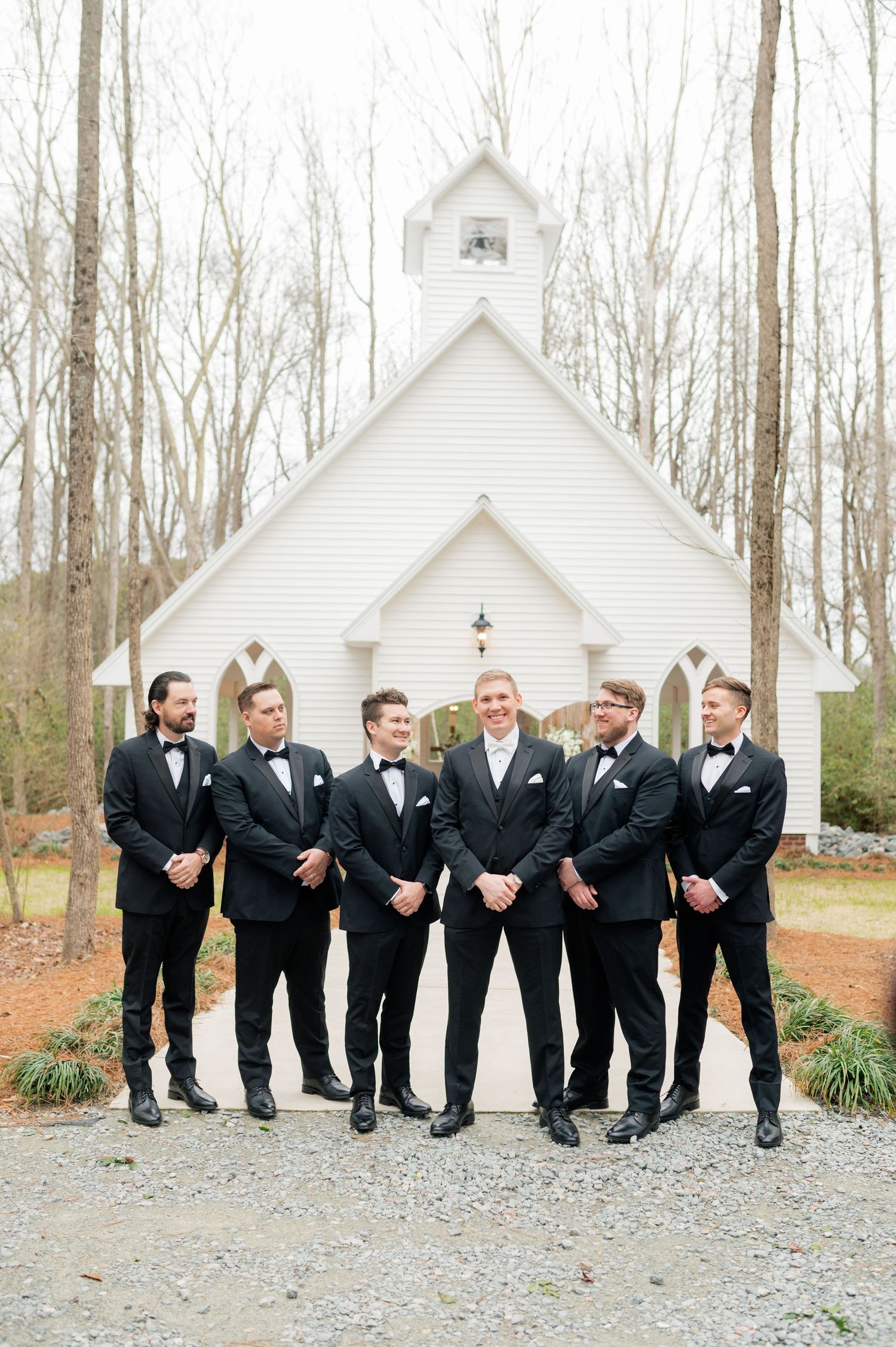 Groomsmen in black tuxedos lined up outside a white chapel in the woods sharing candid moments.