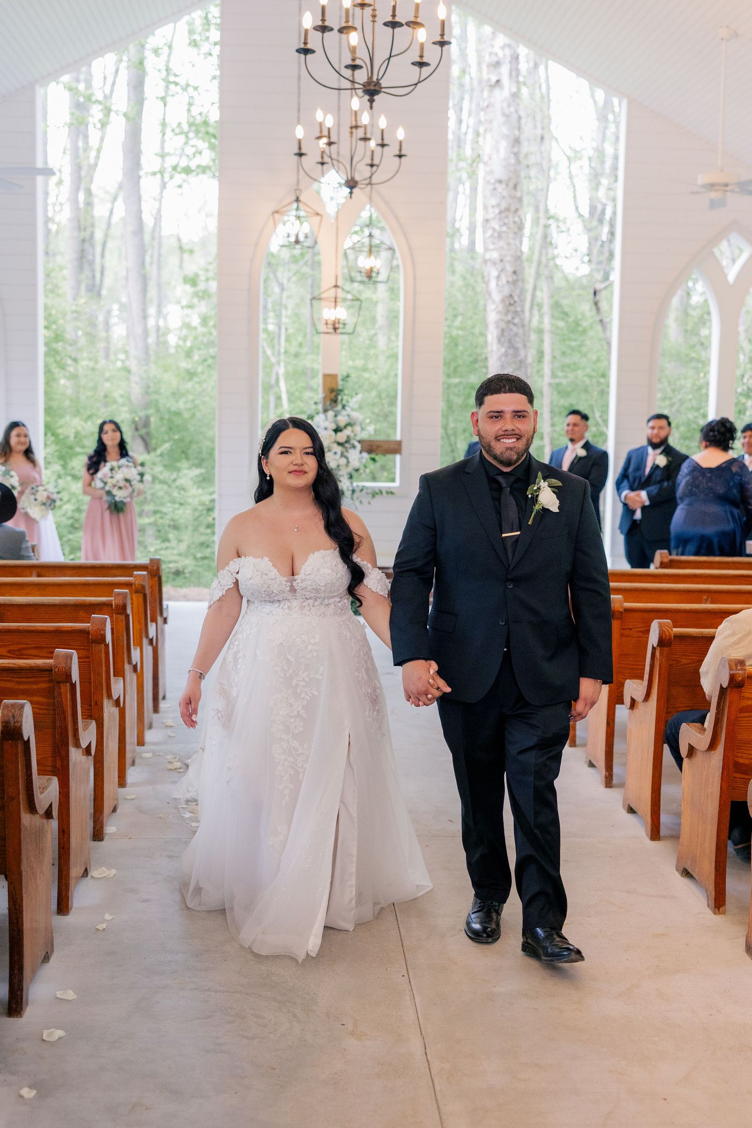 Wedding couple walks down aisle in light-filled chapel with wooden pews and chandeliers.