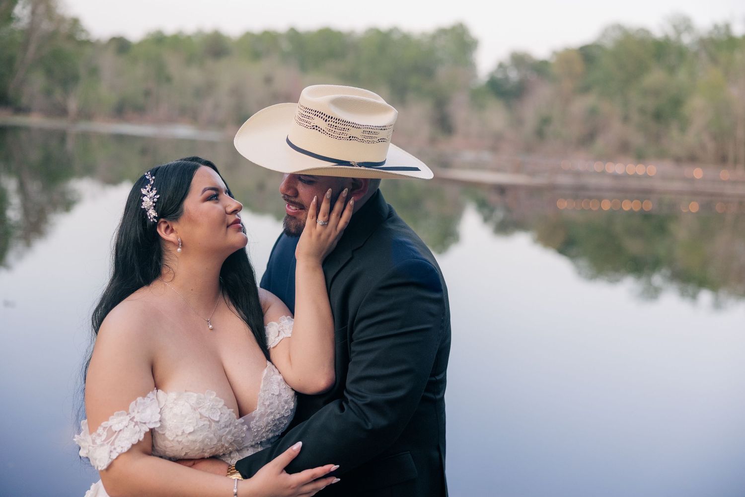 Intimate wedding moment captured by a serene lake with bride in lace dress.