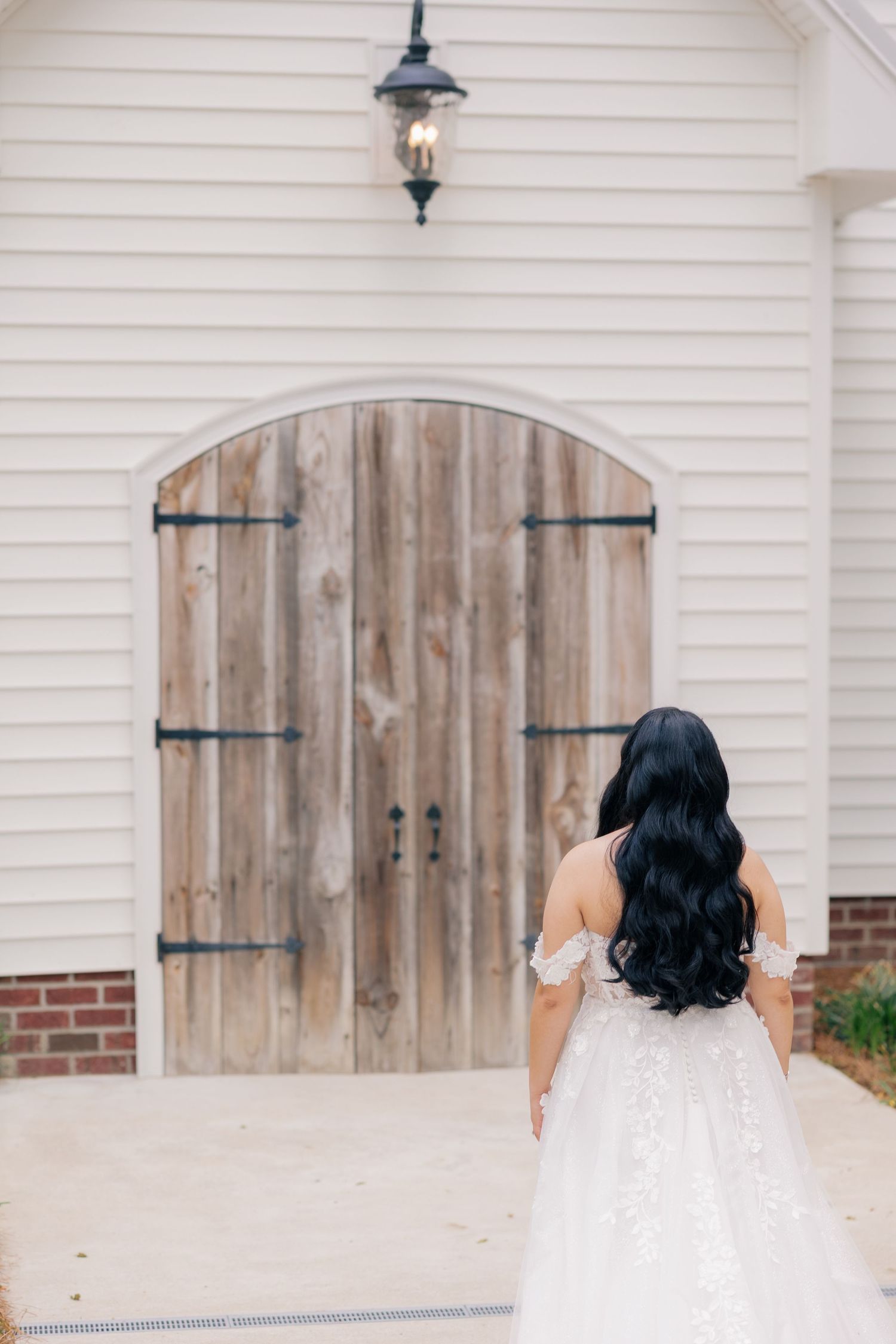 A flowing white wedding dress seen from behind against rustic wooden doors.