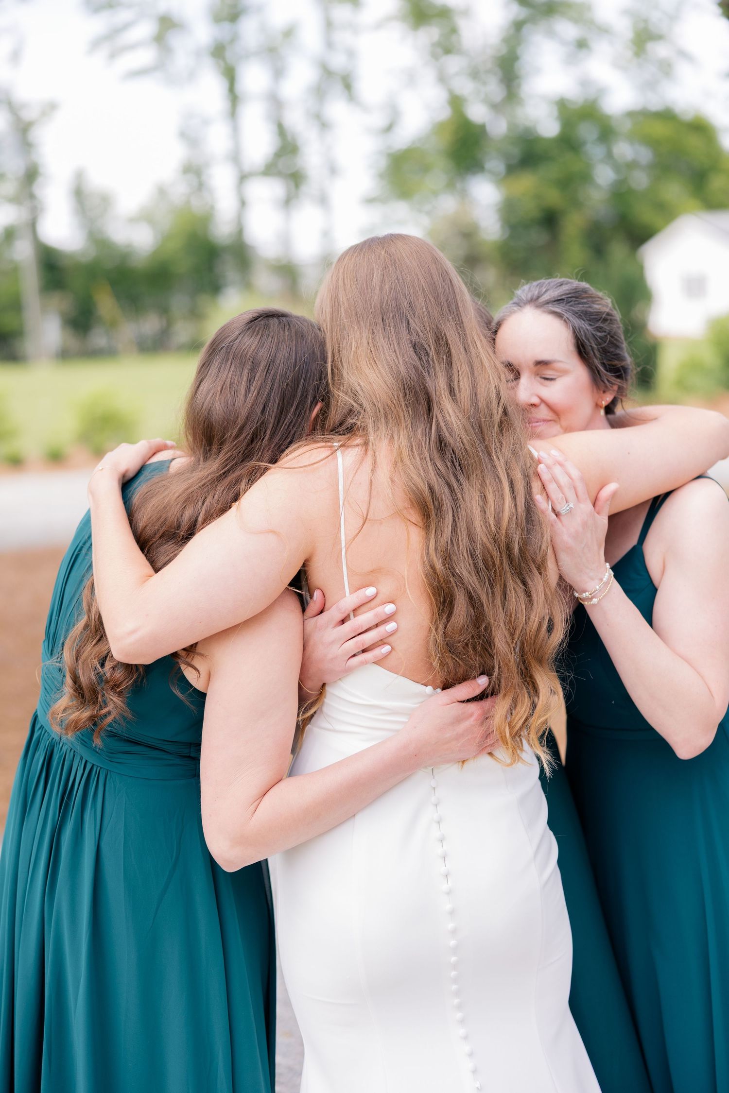 A bride and bridesmaids in teal dresses share emotional embraces outdoors during a wedding celebration.