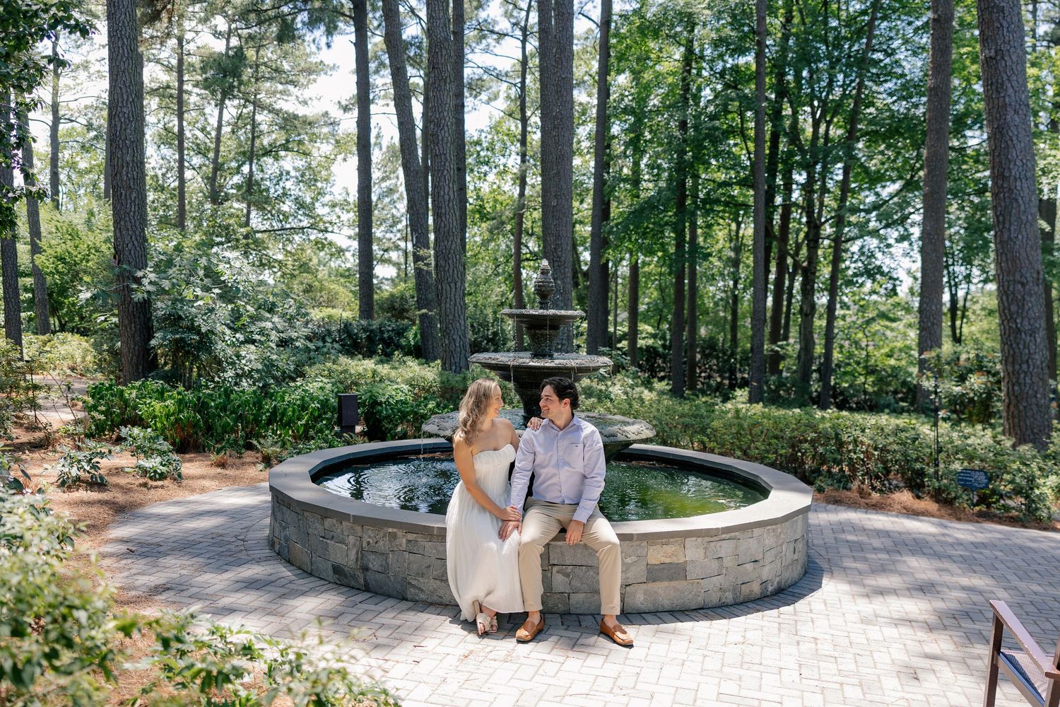 Couple sitting together on stone fountain edge surrounded by tall trees in garden park.